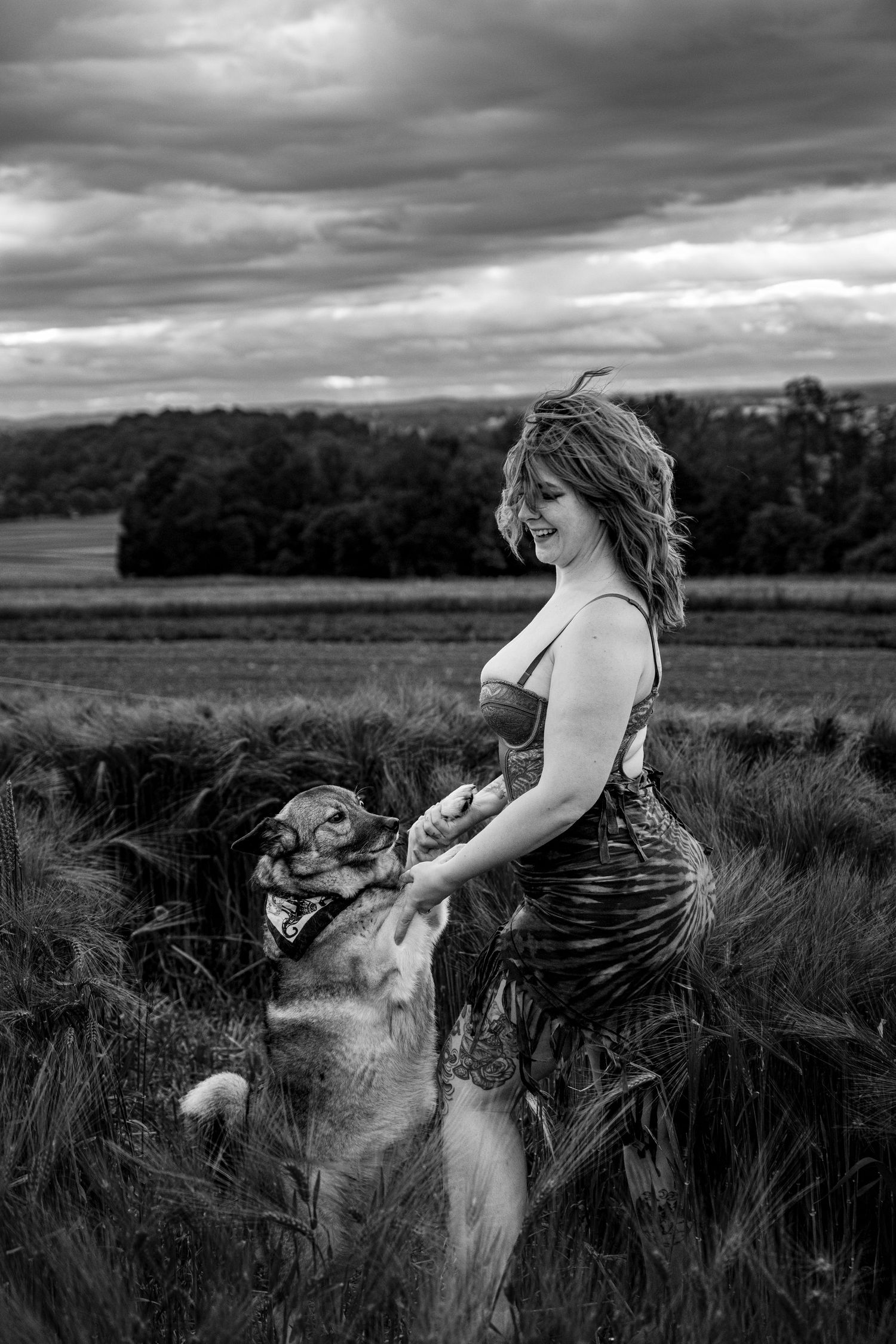 Black and white photo of smiling woman playing with her dog in a dramatic cloudy wheat field.