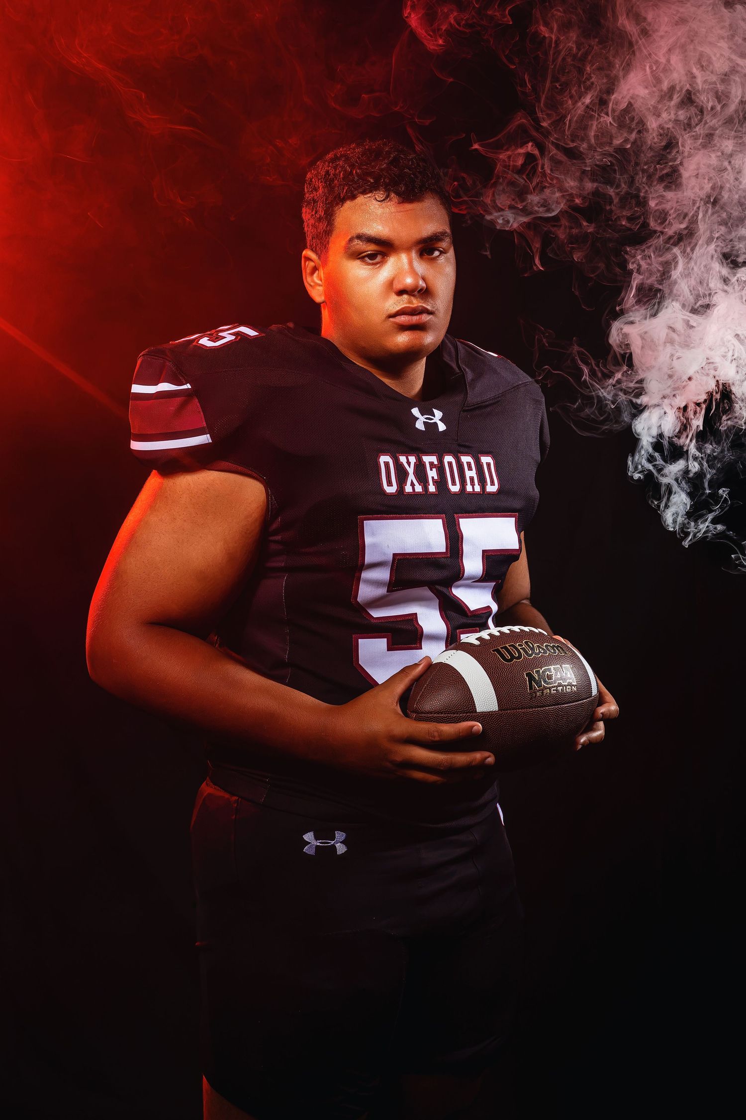 Oxford Chargers #55 football player holds football in moody red smoke studio photo.