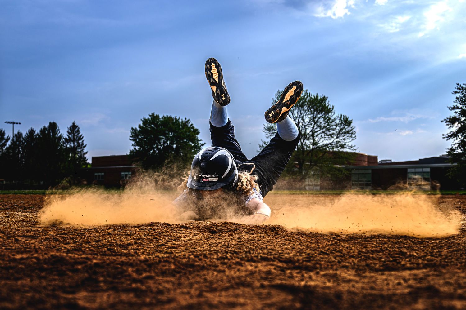 Softball player flips upside down sliding into base with cleats in air and dirt flying.