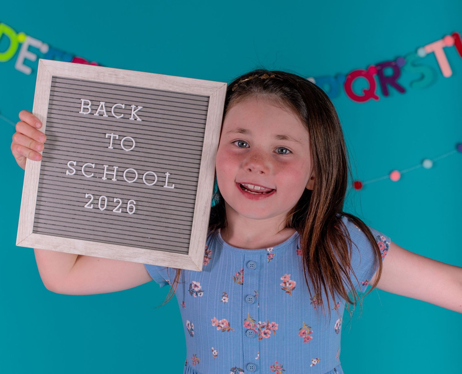 Smiling girl in a floral blue dress holds a Back to School 2026 letter board against a teal background with a colorful felt banner.