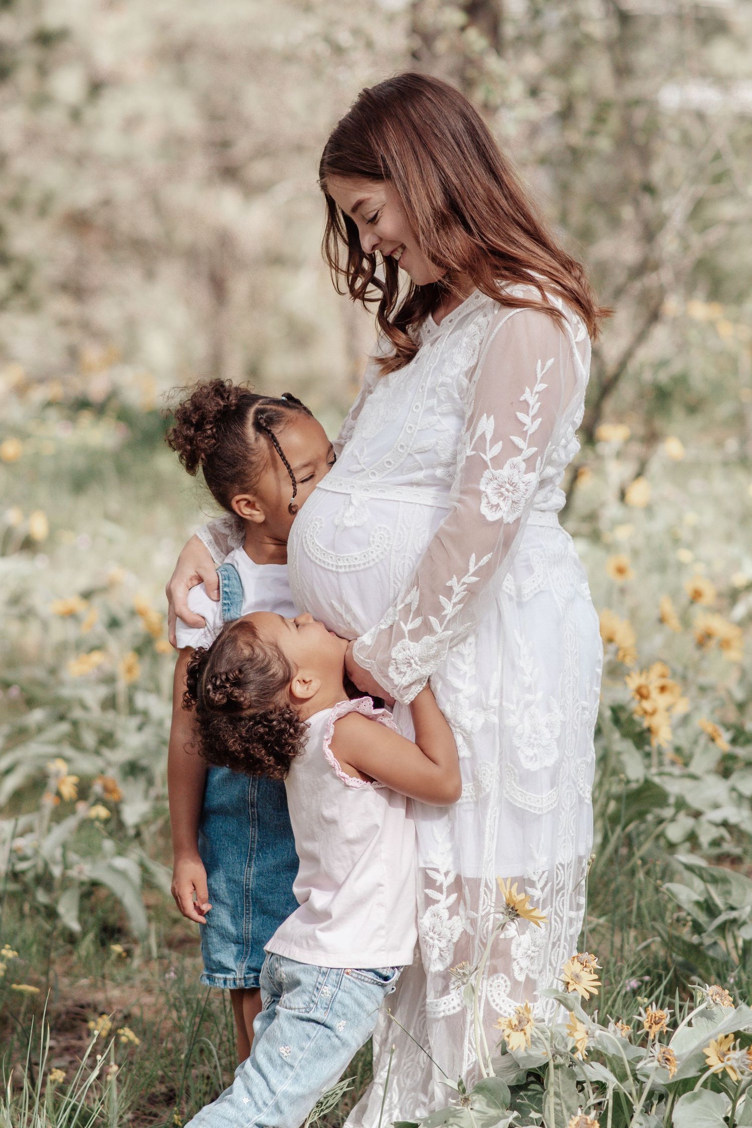 Pregnant woman in a white lace dress smiles down at two young girls hugging her belly in a sunlit field of yellow wildflowers.