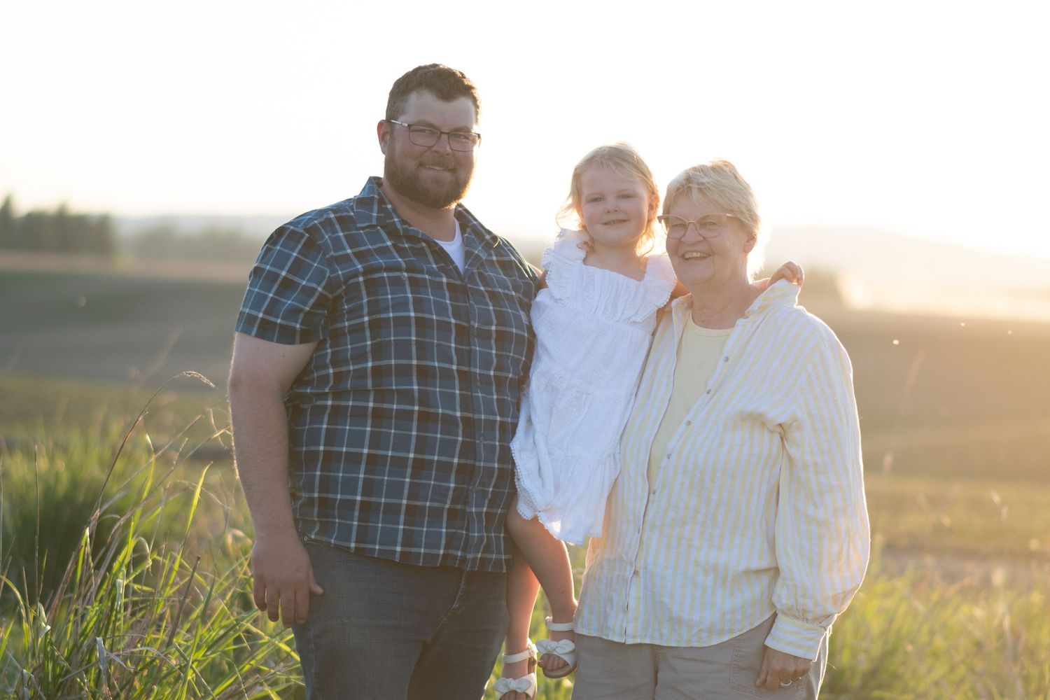Man and older woman pose with young child in golden sunset field family portrait.