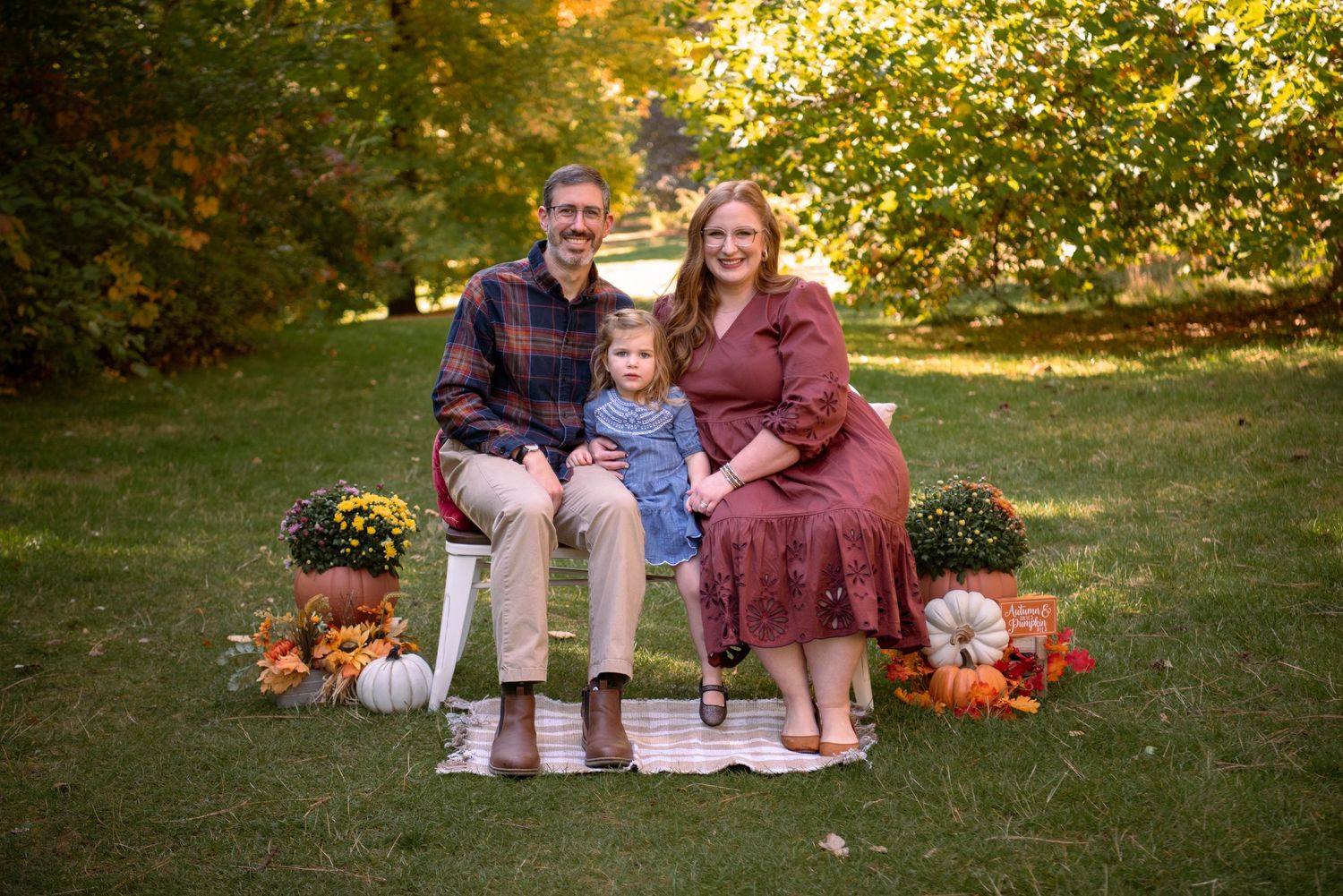 Smiling family of three sits on a white bench in a grassy park surrounded by pumpkins and mums in an autumn portrait.