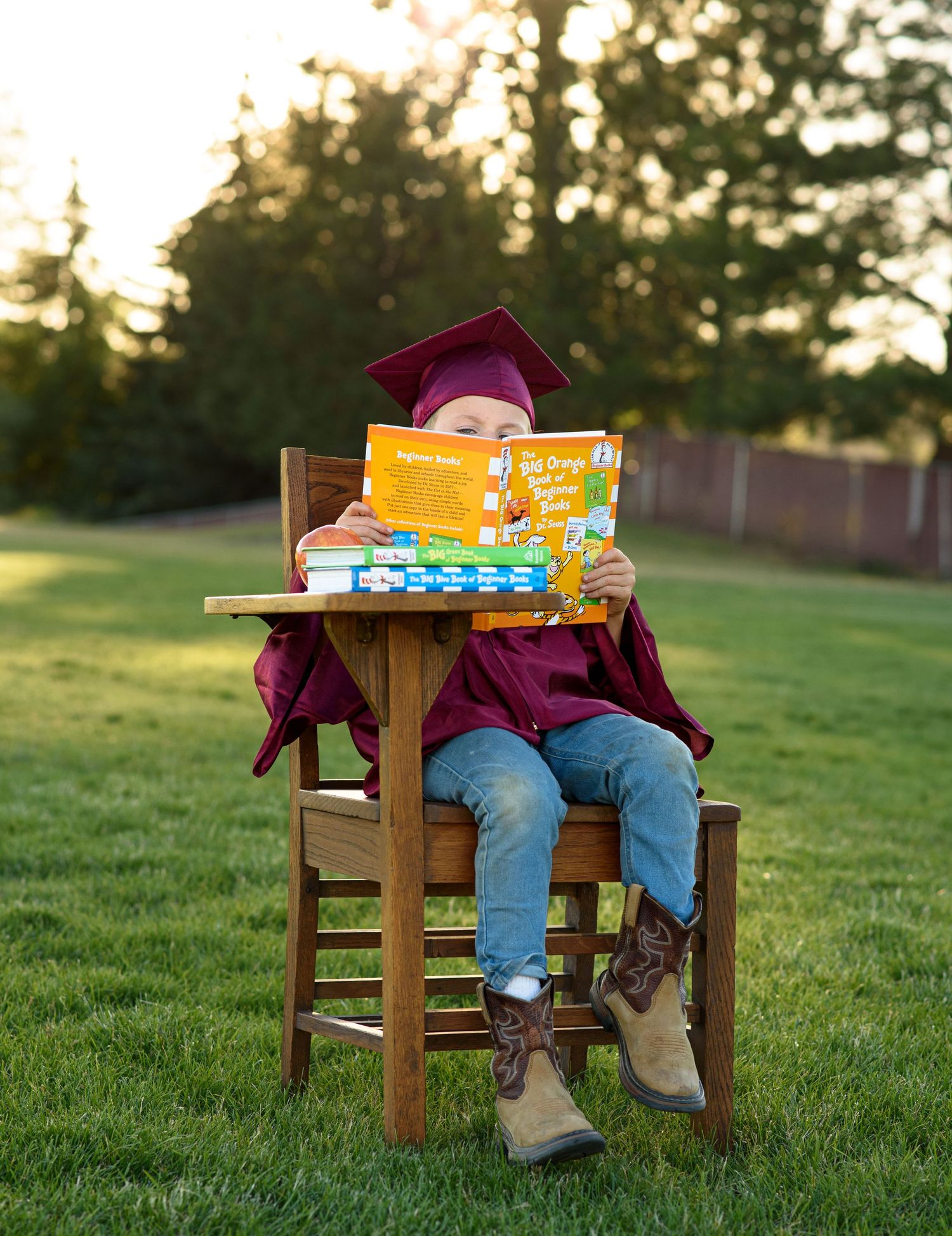Child in a maroon graduation cap and gown sits at a wooden school desk on a grassy lawn, hiding behind a colorful picture book.