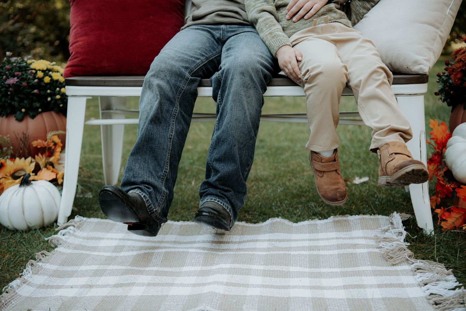 Two people sit on a white bench in jeans and boots, with a plaid blanket, pumpkins and fall flowers on the grass for an autumn photo.
