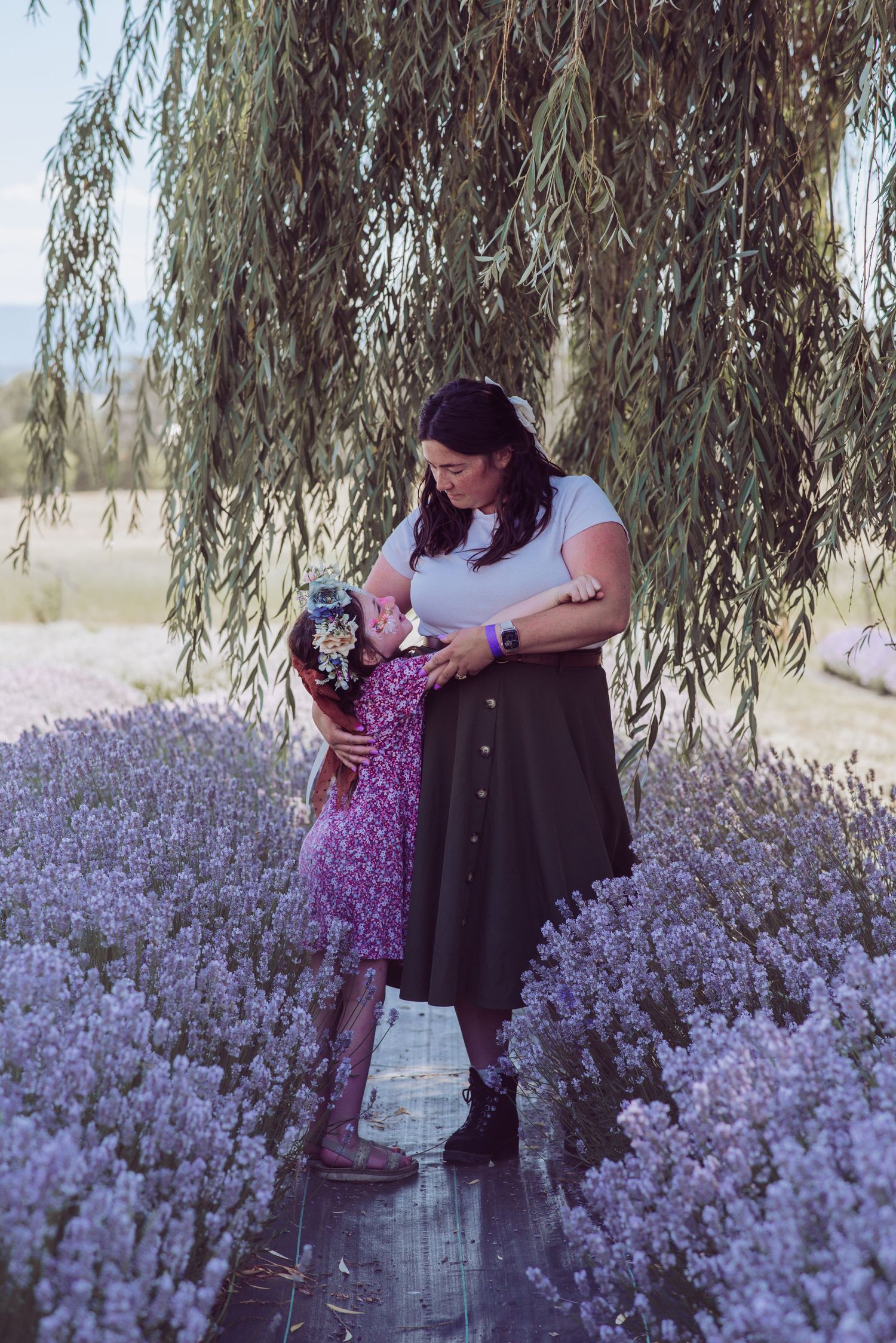 A mother and young daughter embracing in a beautiful lavender field under a weeping willow tree.