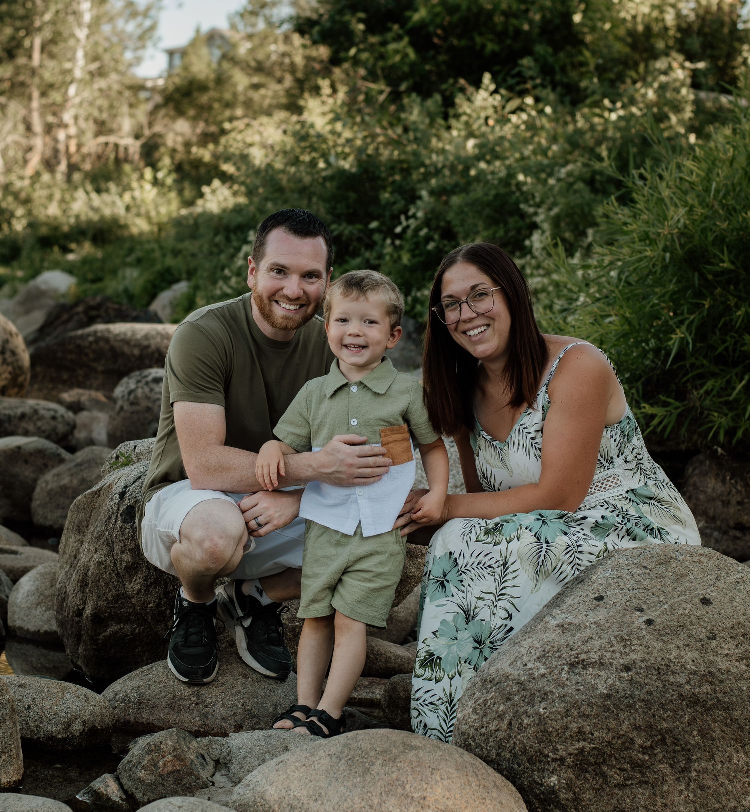 A happy family of three poses on large rocks surrounded by lush green nature.