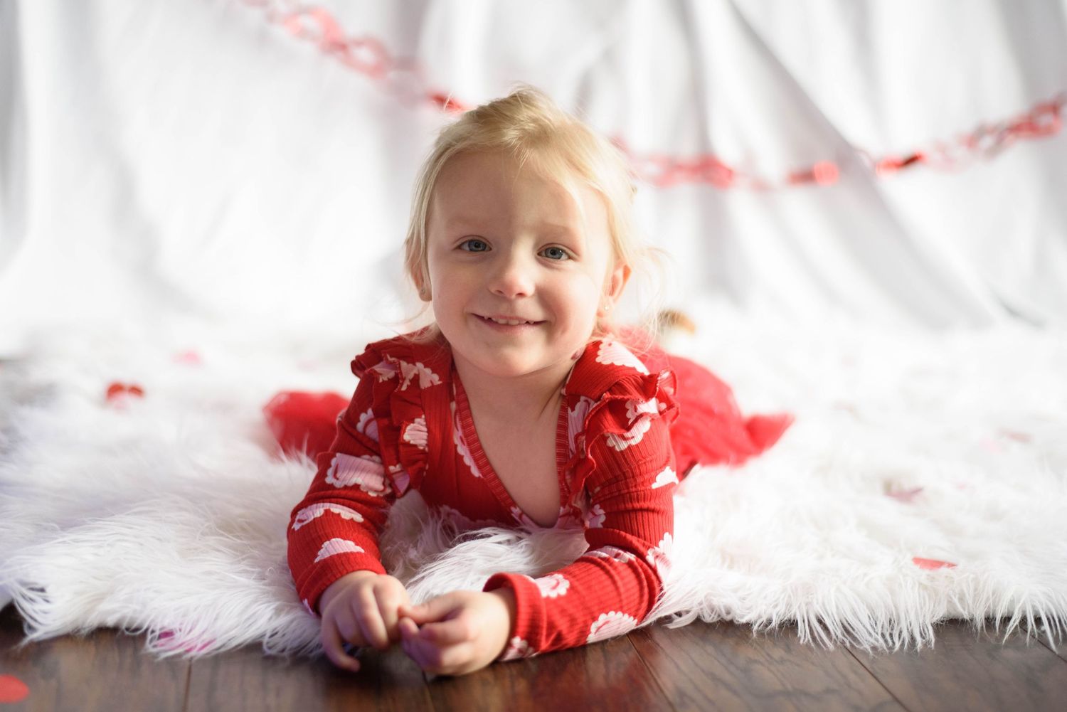 Smiling toddler in red holiday pajamas lies on a white fur rug with a heart garland and scattered petals in the background.