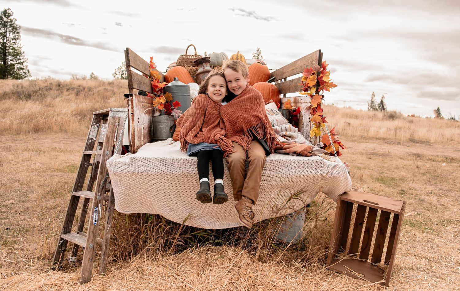 Two children snuggle together on a blanket-covered truck bed decorated with pumpkins, autumn leaves, and hay in a golden fall field.