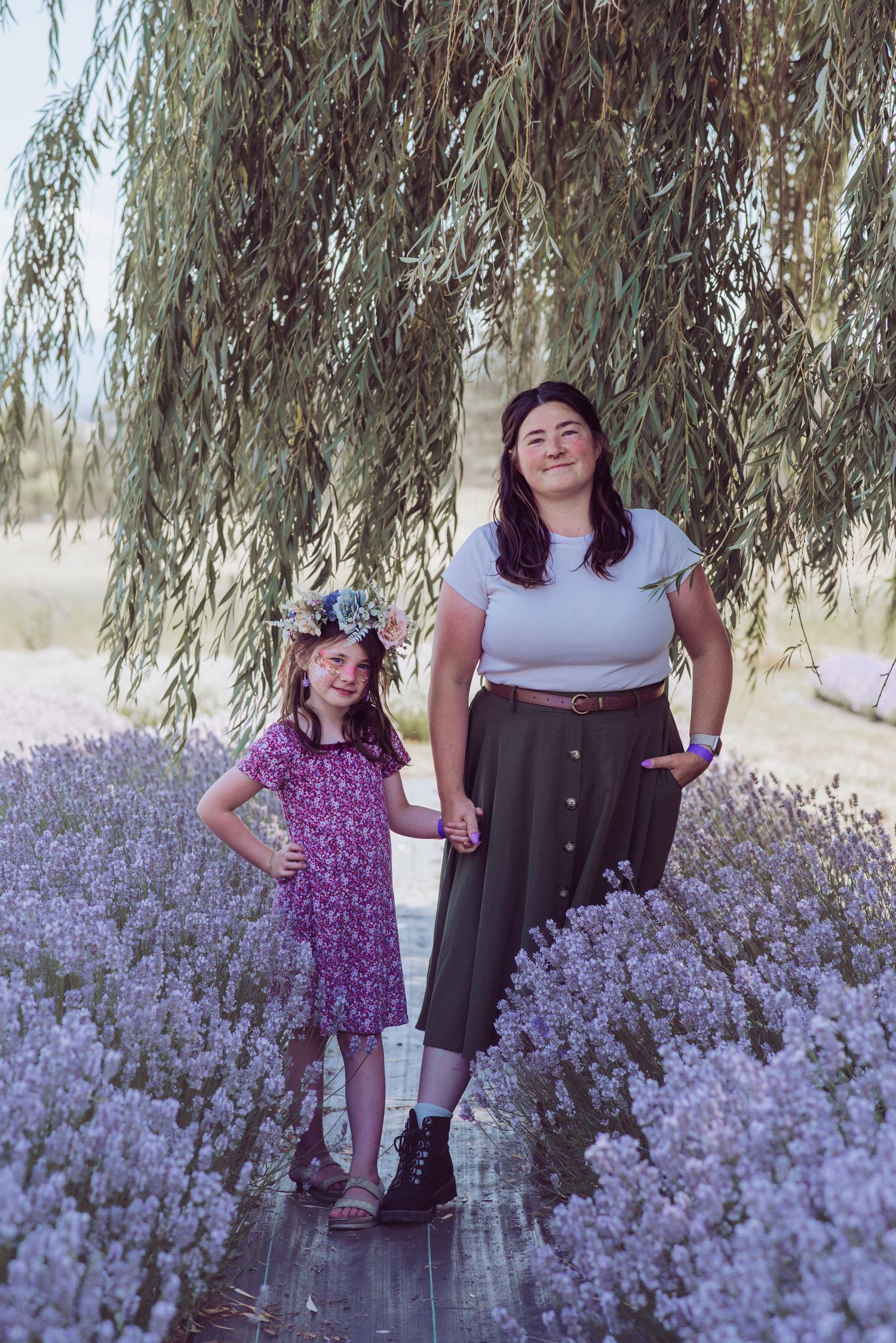 A woman and a young girl in a flower crown hold hands on a wooden path between lavender rows beneath a weeping willow tree.