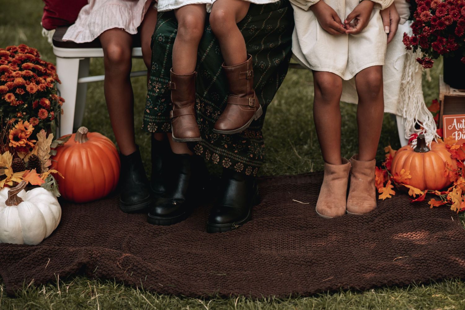 Three children in fall boots sit with pumpkins and autumn mums in seasonal photo.