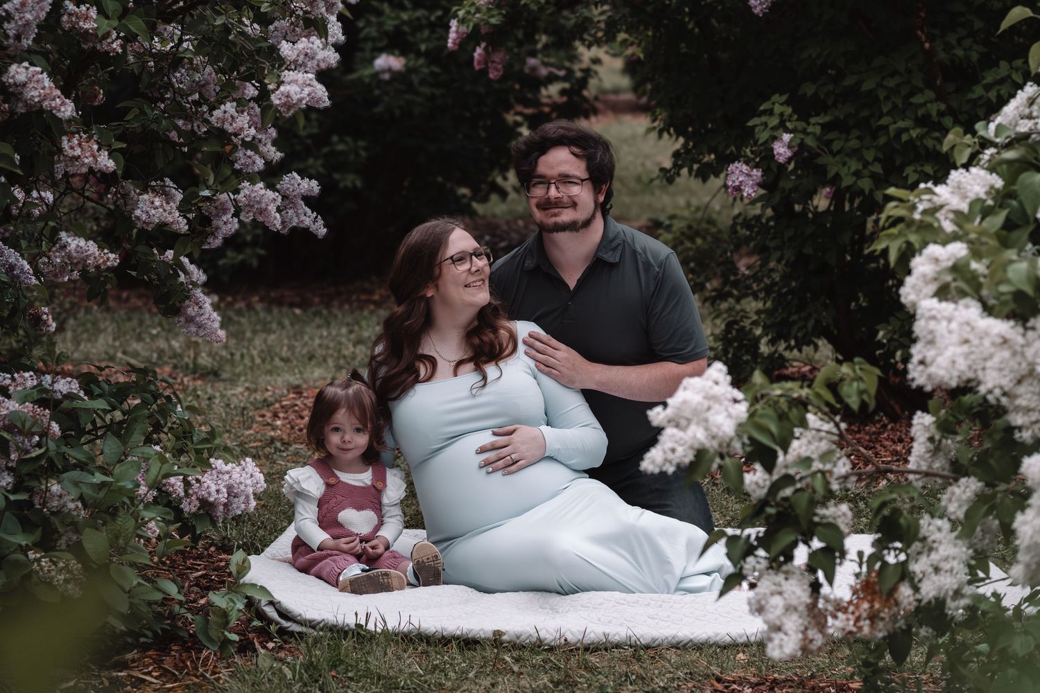 A pregnant woman poses with her partner and young daughter among blooming lilac bushes.