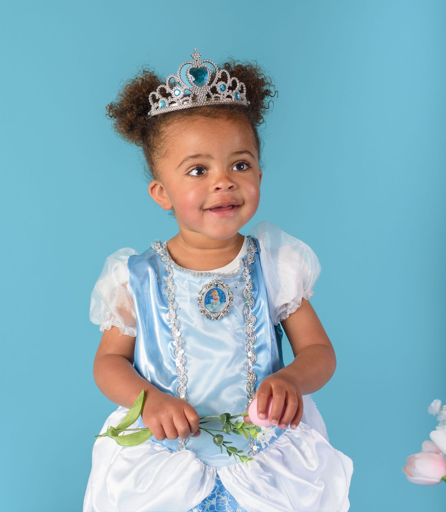 An adorable toddler girl in a Cinderella dress and tiara smiles against a bright blue backdrop.