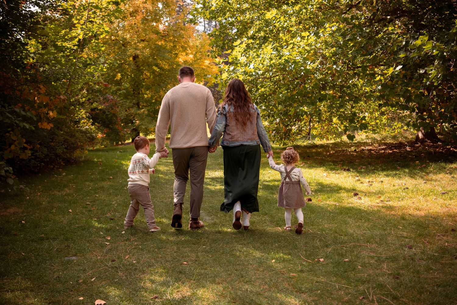Family of four walking hand in hand away from the camera across a sunny grassy park lined with colorful autumn trees.