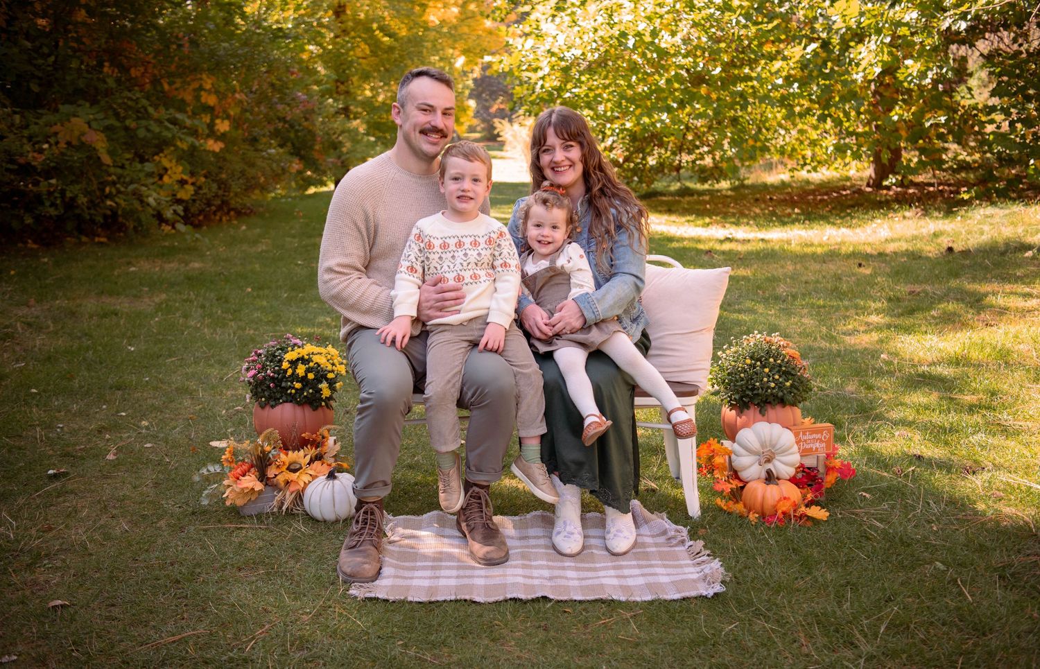 Smiling family of four sitting on a white bench with two young children on a striped blanket surrounded by pumpkins and mums in autumn.