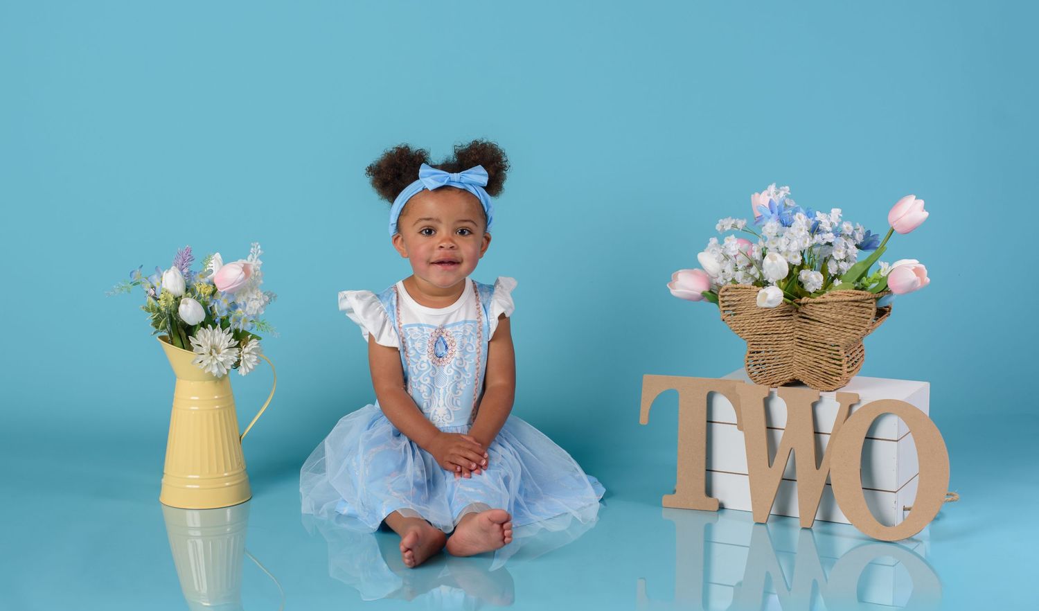 Toddler girl in blue dress sits beside TWO sign and flowers for birthday portraits.