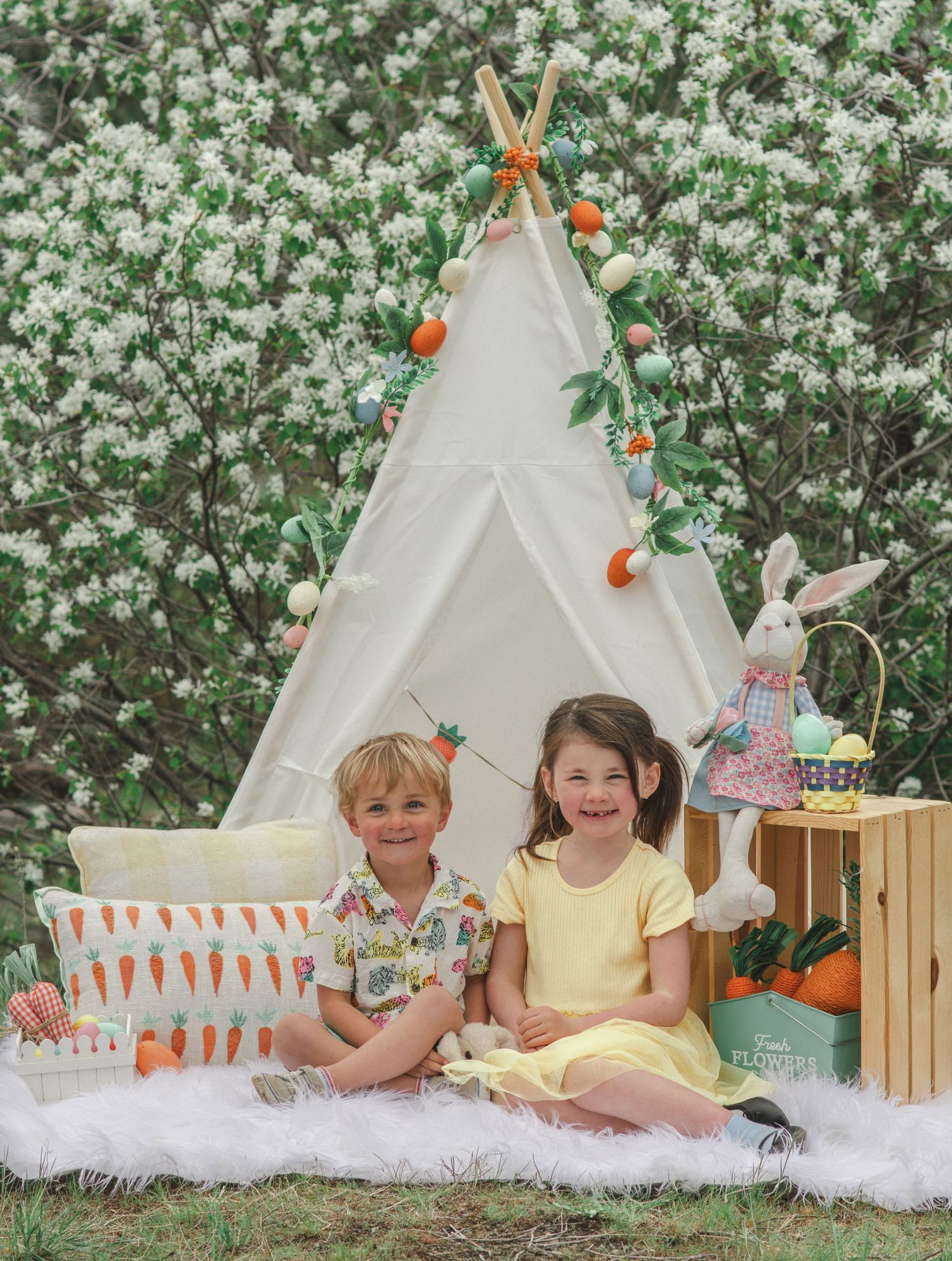 Two smiling children sit on a white fur rug beside a teepee with carrot garland, an Easter bunny and basket, and blooming white trees.