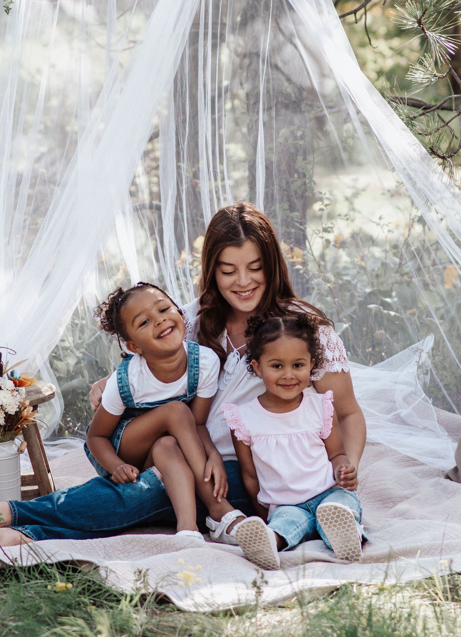 A smiling woman sits on a blanket with two laughing little girls under a white canopy in a sunny, grassy outdoor setting.