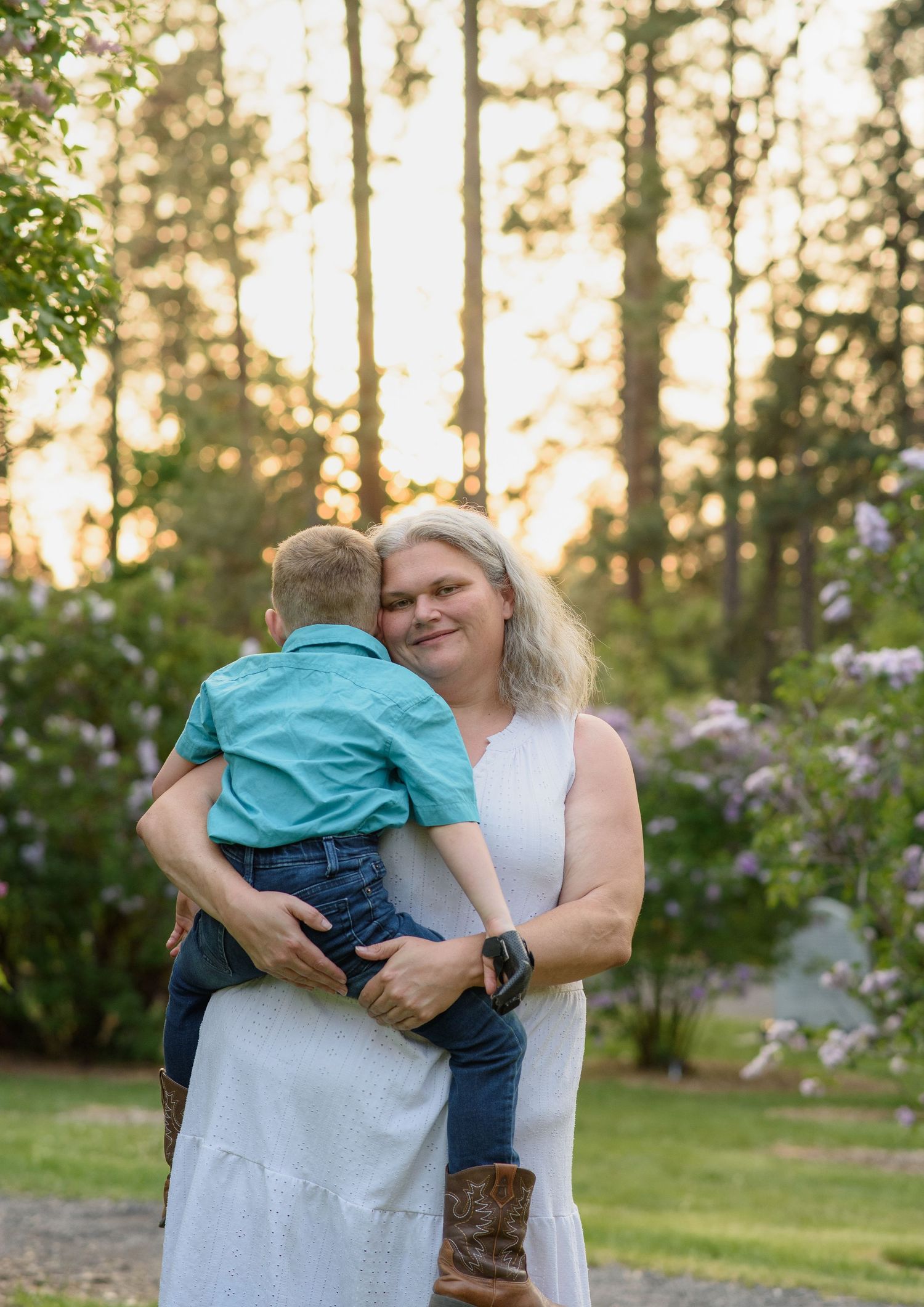 Woman holding young boy in sunny outdoor setting with pine trees and flowers.