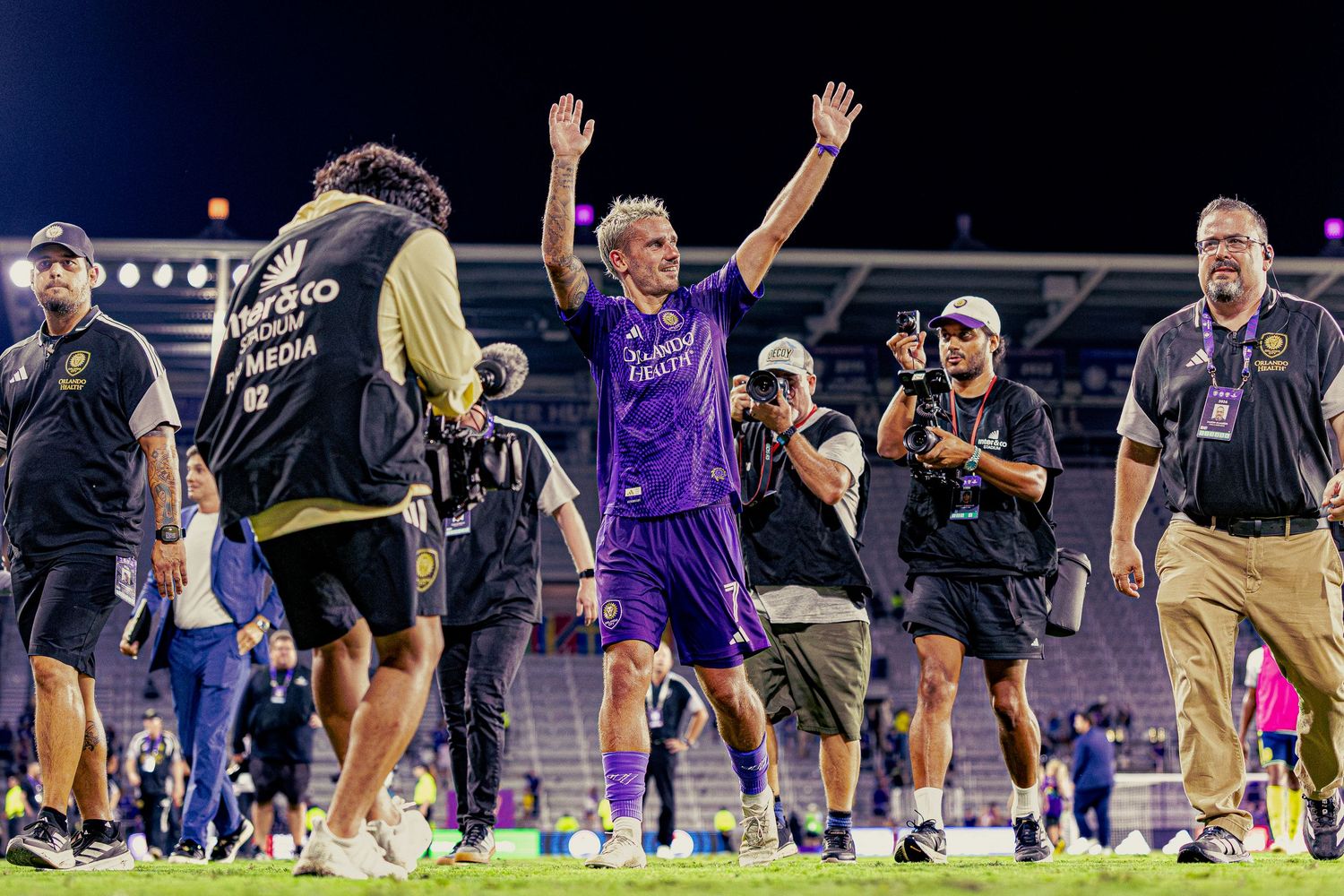 Soccer player in purple jersey waves to crowd on field at night, surrounded by team staff in gold uniforms.