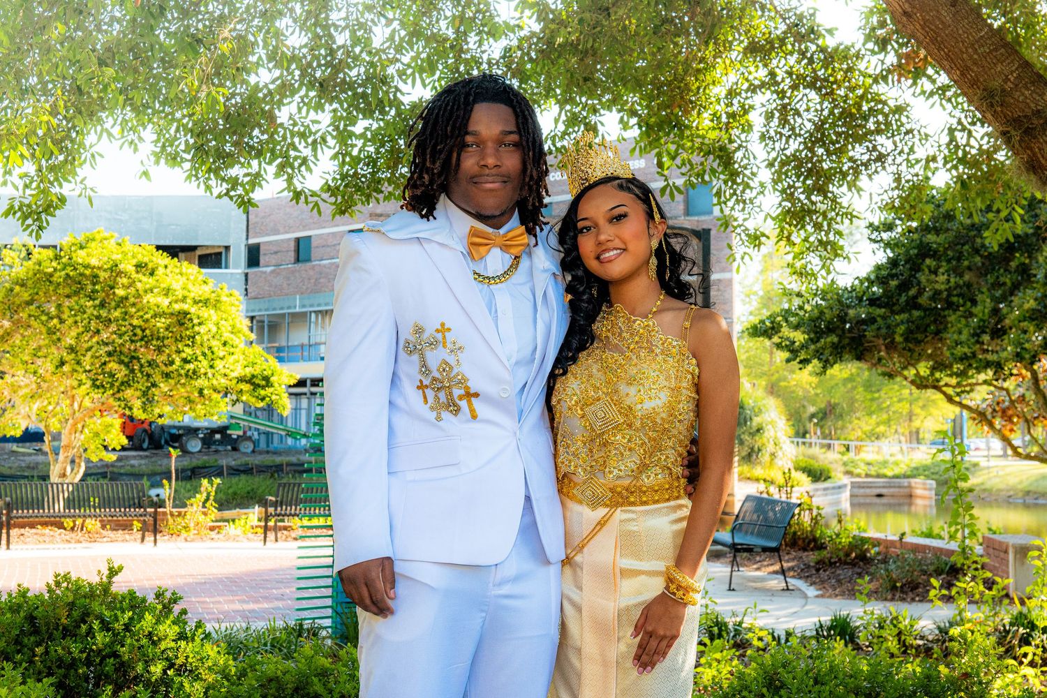 Couple in matching gold and white formal attire posing outdoors at prom event surrounded by green trees.