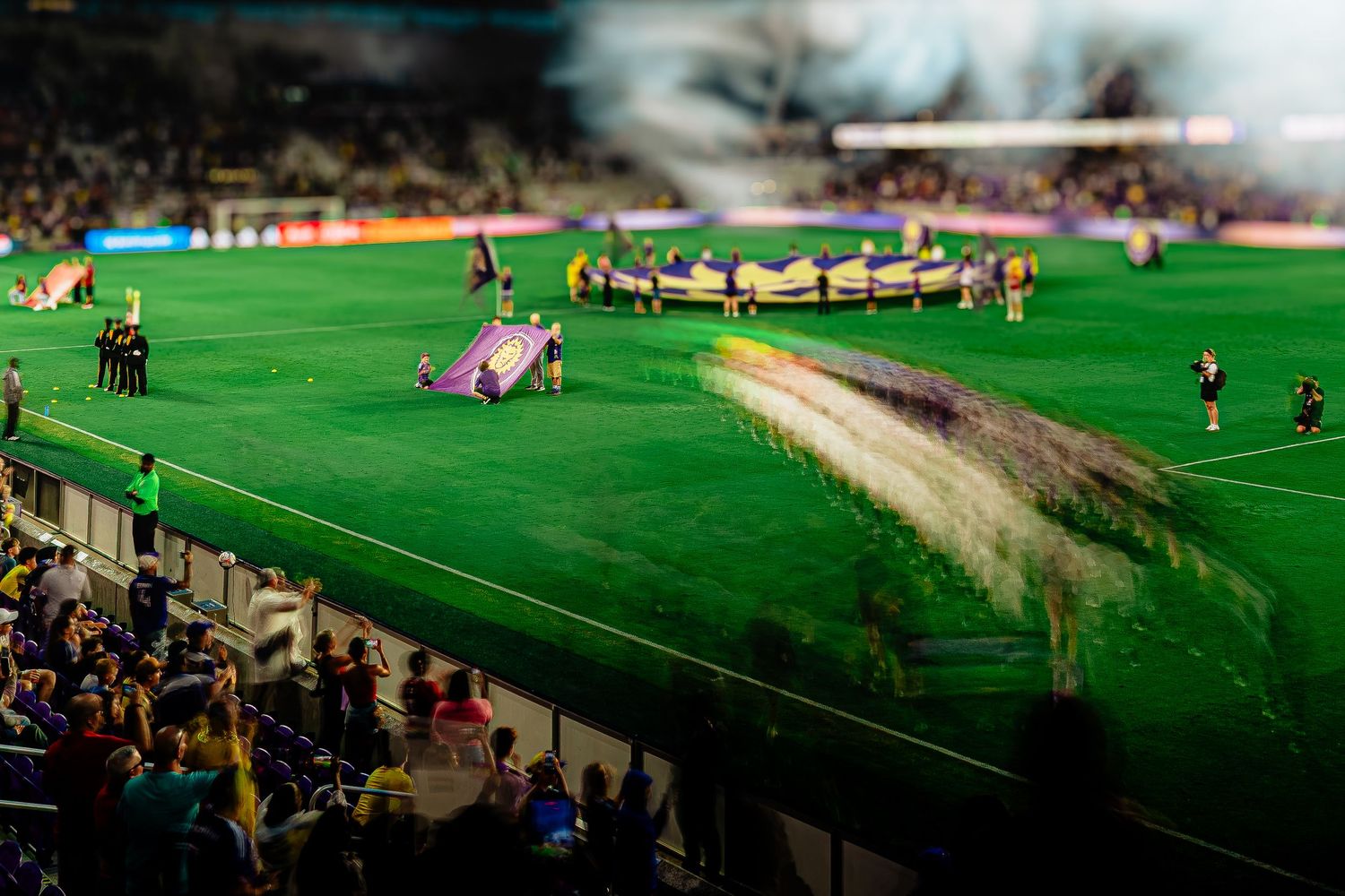 Soccer players unfurl a large banner on a vibrant green stadium field during a nighttime match ceremony.