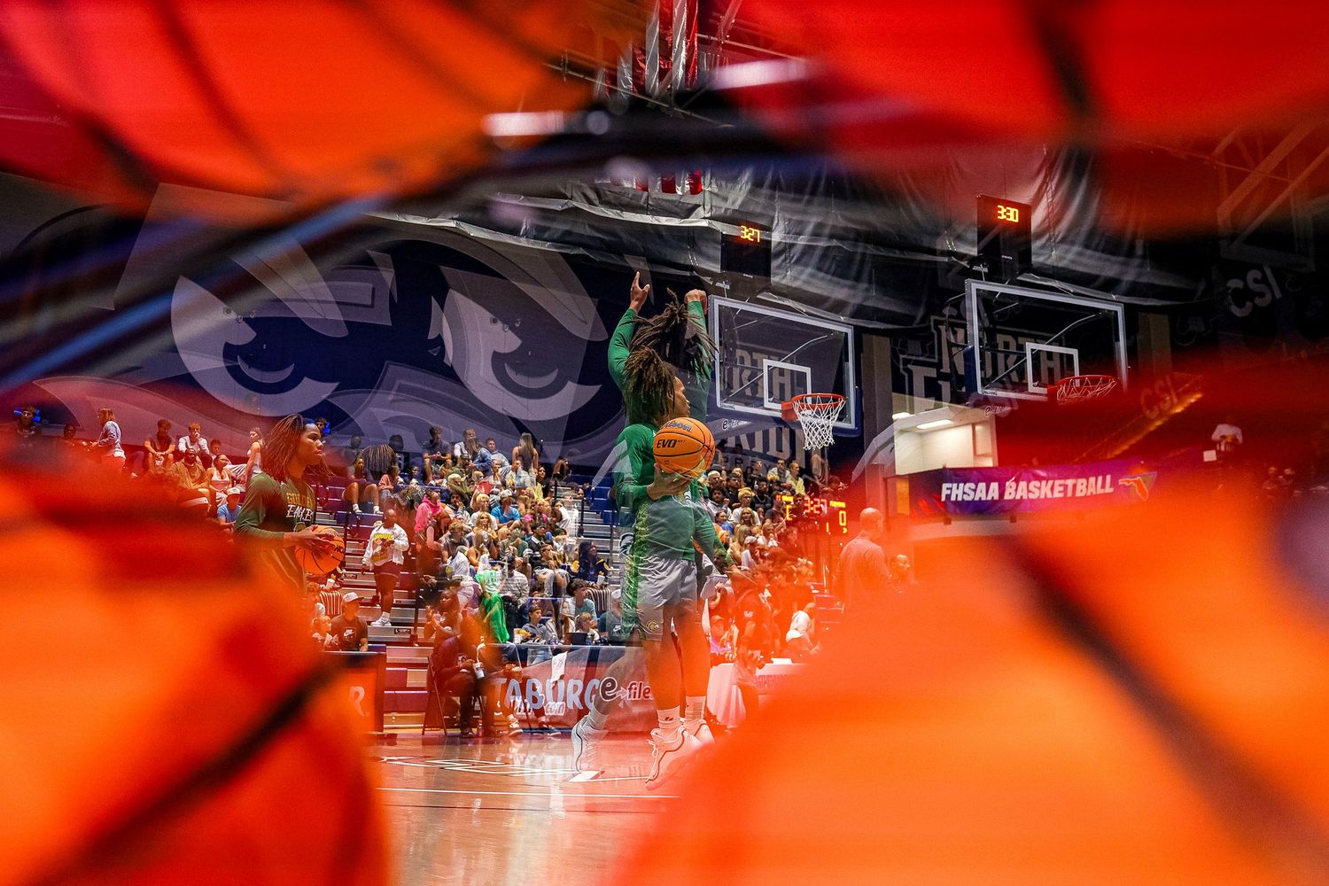 Basketball game viewed through net with crowd and court visible in dramatic red lighting.