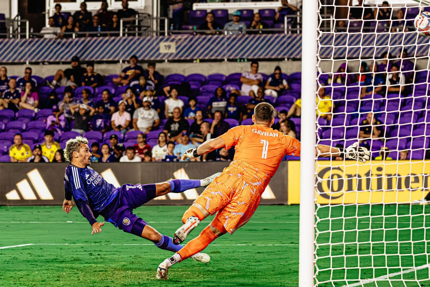 Soccer player scores goal past diving goalkeeper in MLS match at purple-themed stadium.