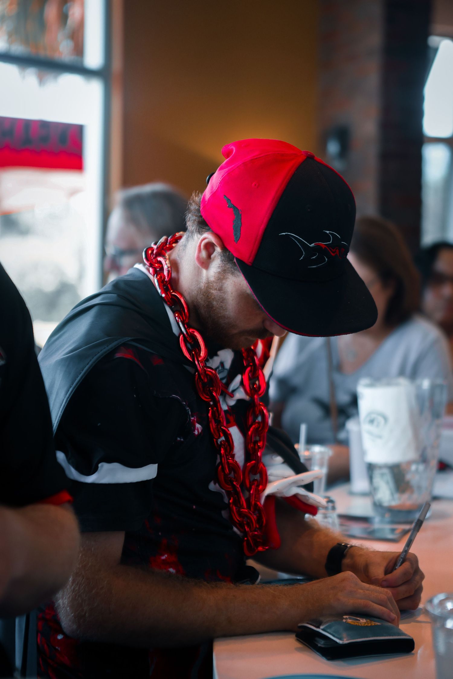 Person wearing red cap and black hoodie looking down at mobile device in restaurant setting.