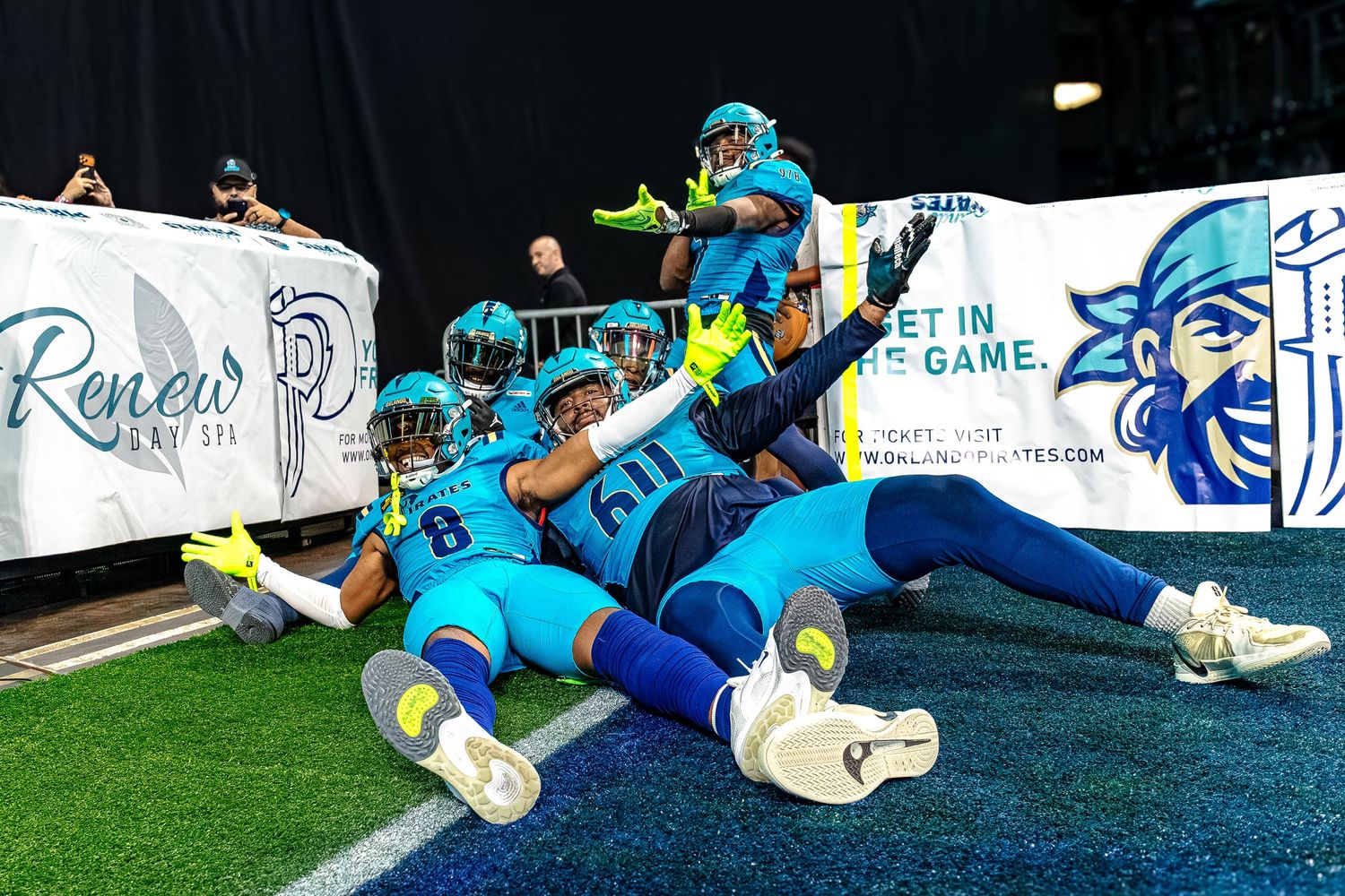 Football players in teal uniforms celebrate a touchdown near the end zone during an indoor arena football game.