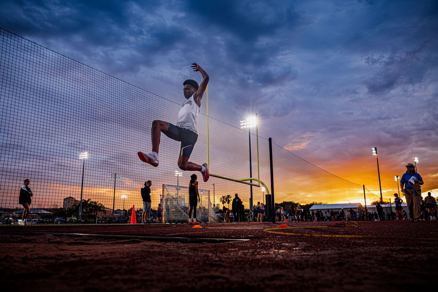 Athlete performing high jump silhouetted against dramatic sunset sky with stadium lights glowing in background.