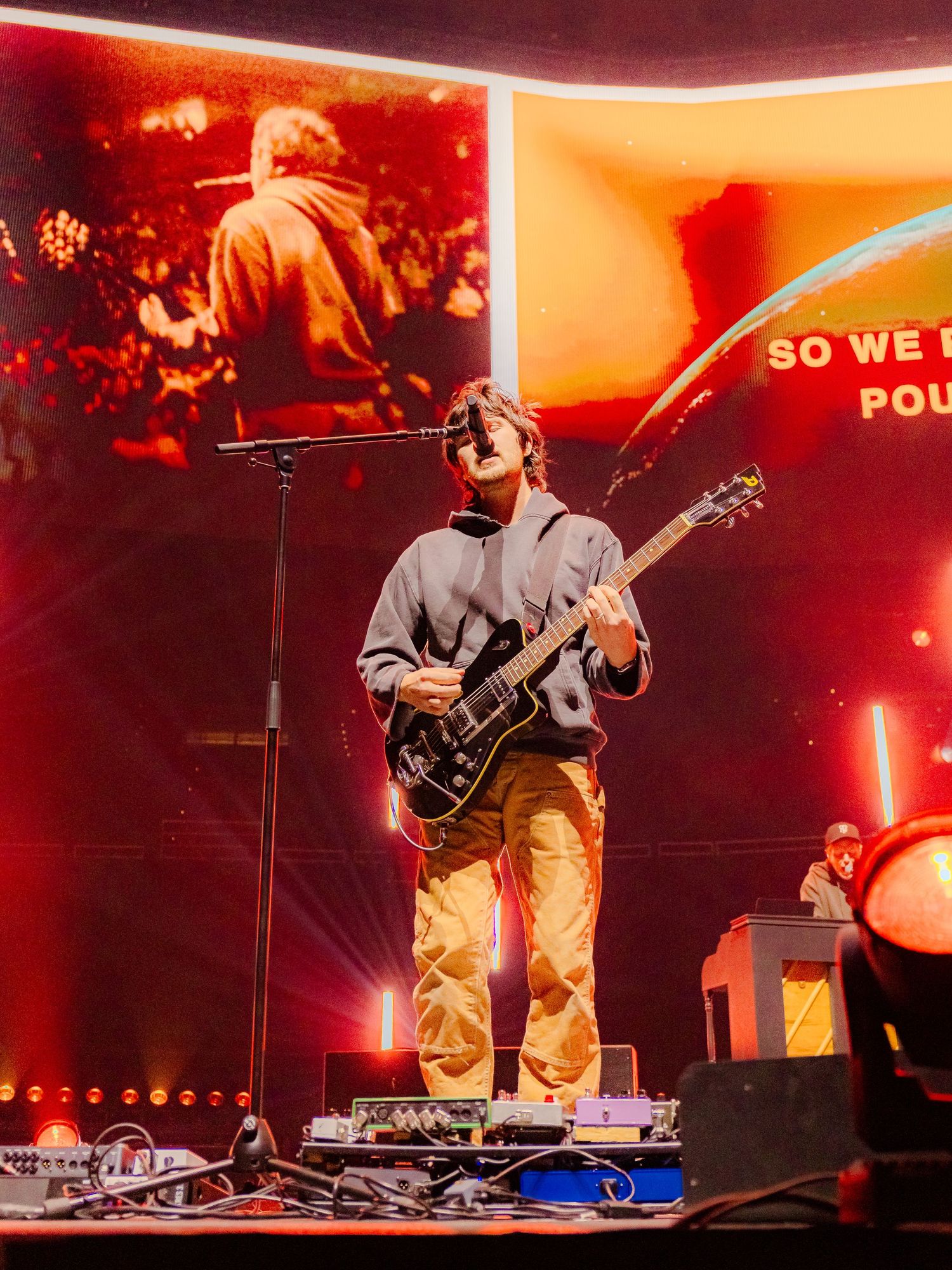 Musician playing bass guitar on stage with red and orange lighting and video screens.