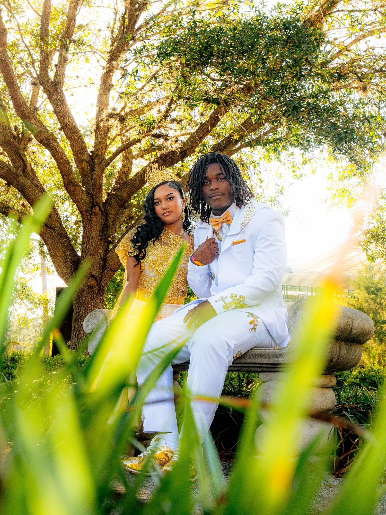 A couple in matching gold and white prom outfits poses on a bench under a tree in a lush garden setting.