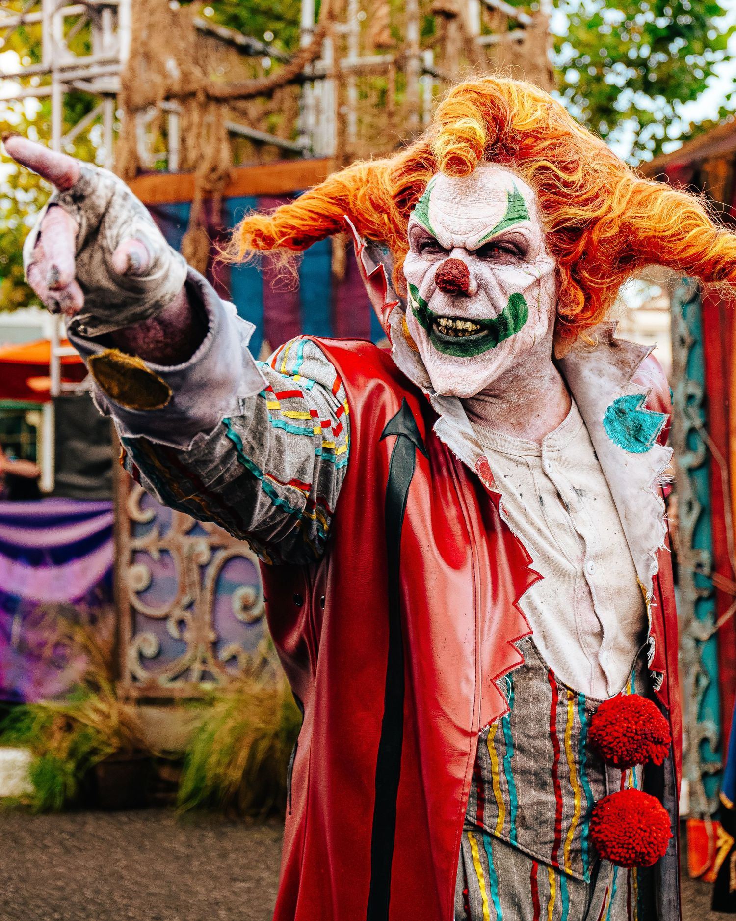 Scary clown costume with orange hair, white face paint, and red cape at a Halloween horror attraction.