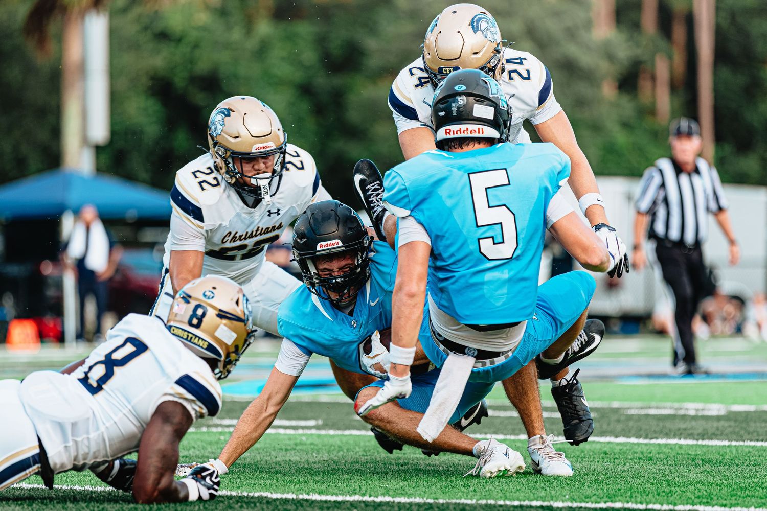 High school football players in teal and navy uniforms collide during an intense tackle play on a green field.