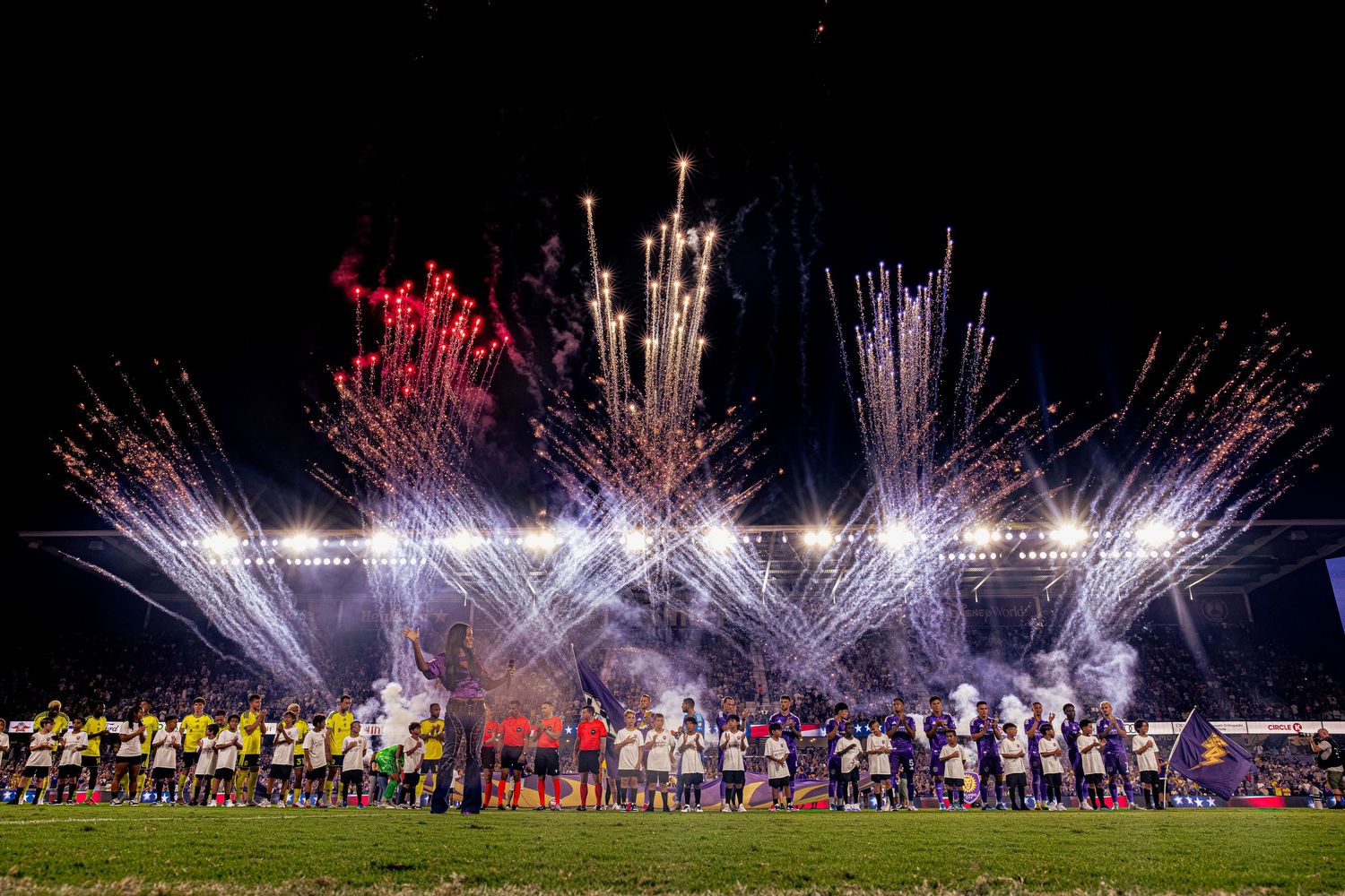 Soccer teams line up on field with spectacular fireworks display lighting up the night sky during a celebratory match event.
