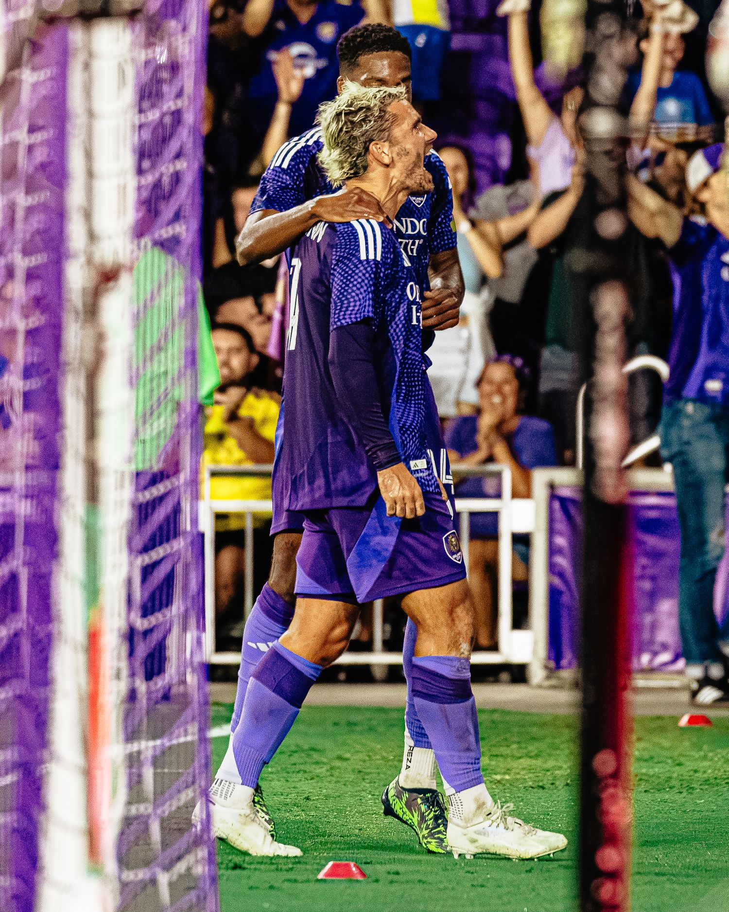 Two Orlando City SC players celebrate a goal in purple uniforms at a night MLS soccer match.