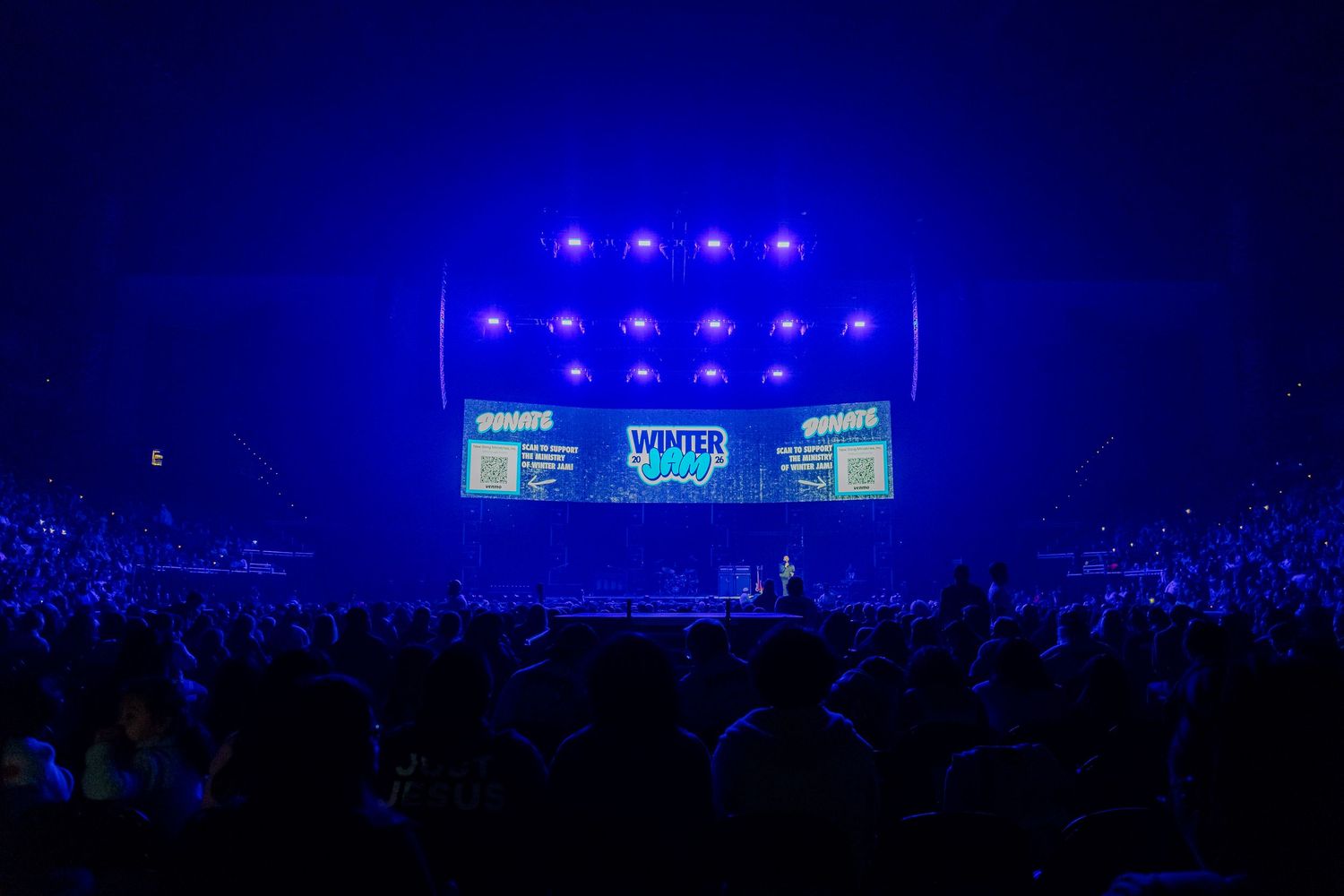 Large concert venue with blue stage lighting and crowd silhouettes against dark background.