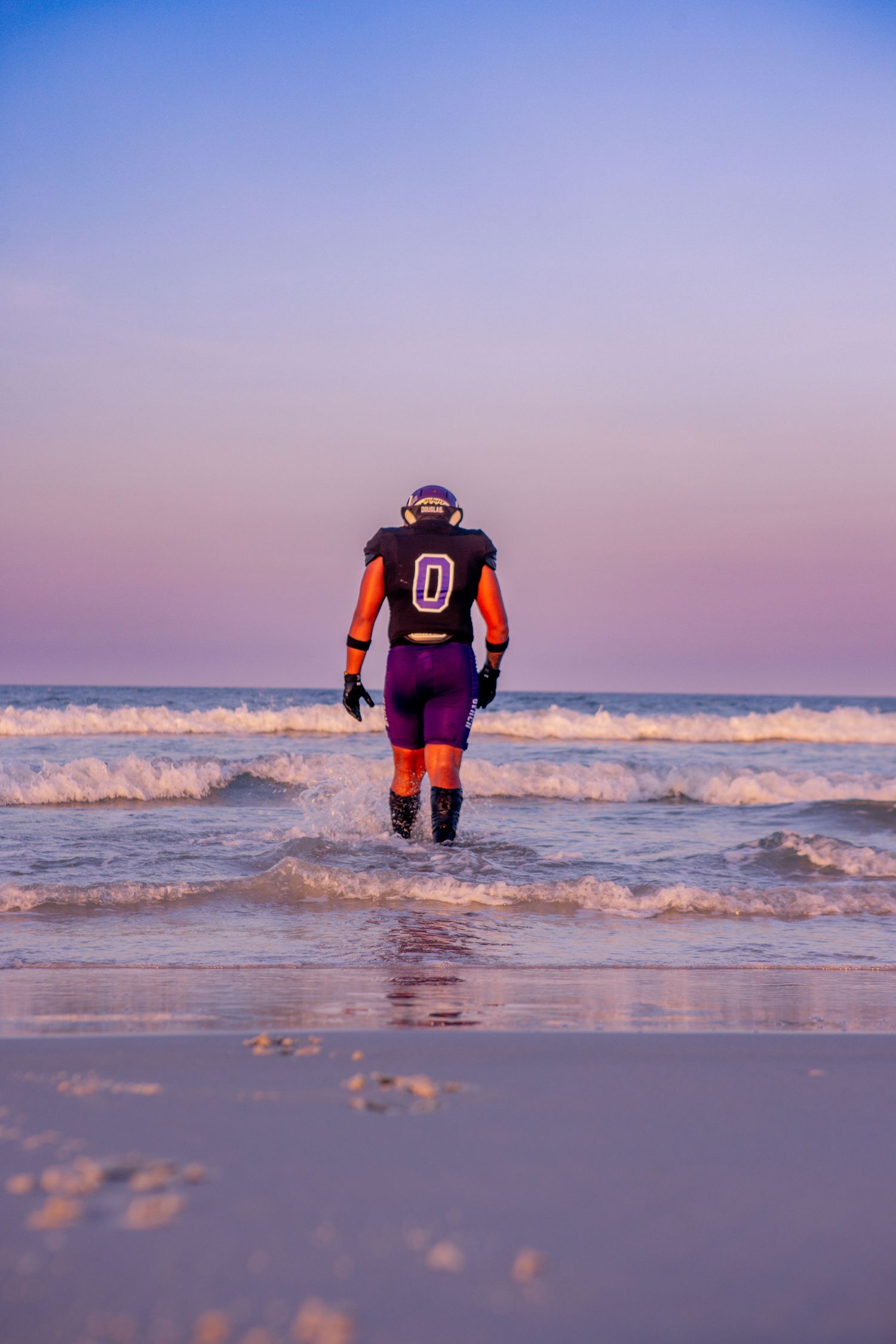 Person wearing team jersey wades into ocean waves at sunset.