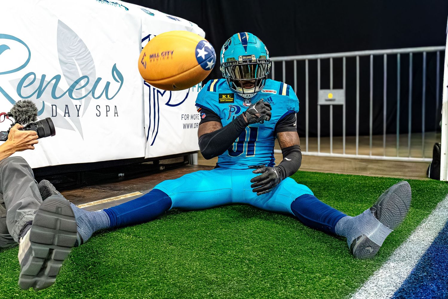 Football player in teal uniform does splits pose on field near Renew Day Spa banner holding football.