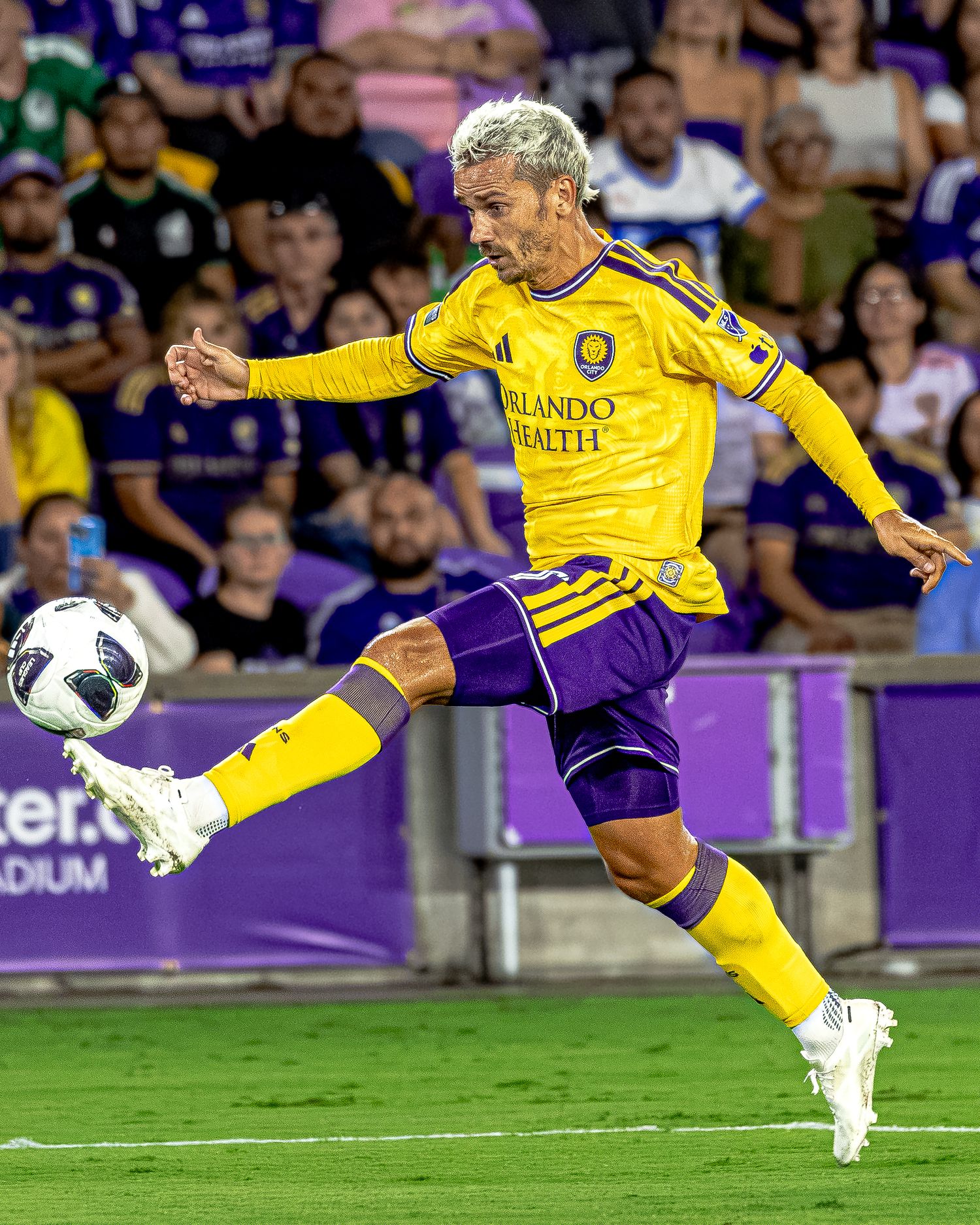 Orlando City SC player in yellow jersey controls soccer ball mid-air during MLS match at purple-themed stadium.