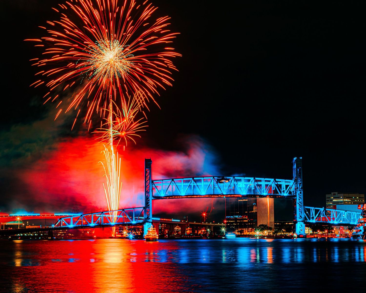 Fireworks display illuminates night sky over blue-lit bridge structure.