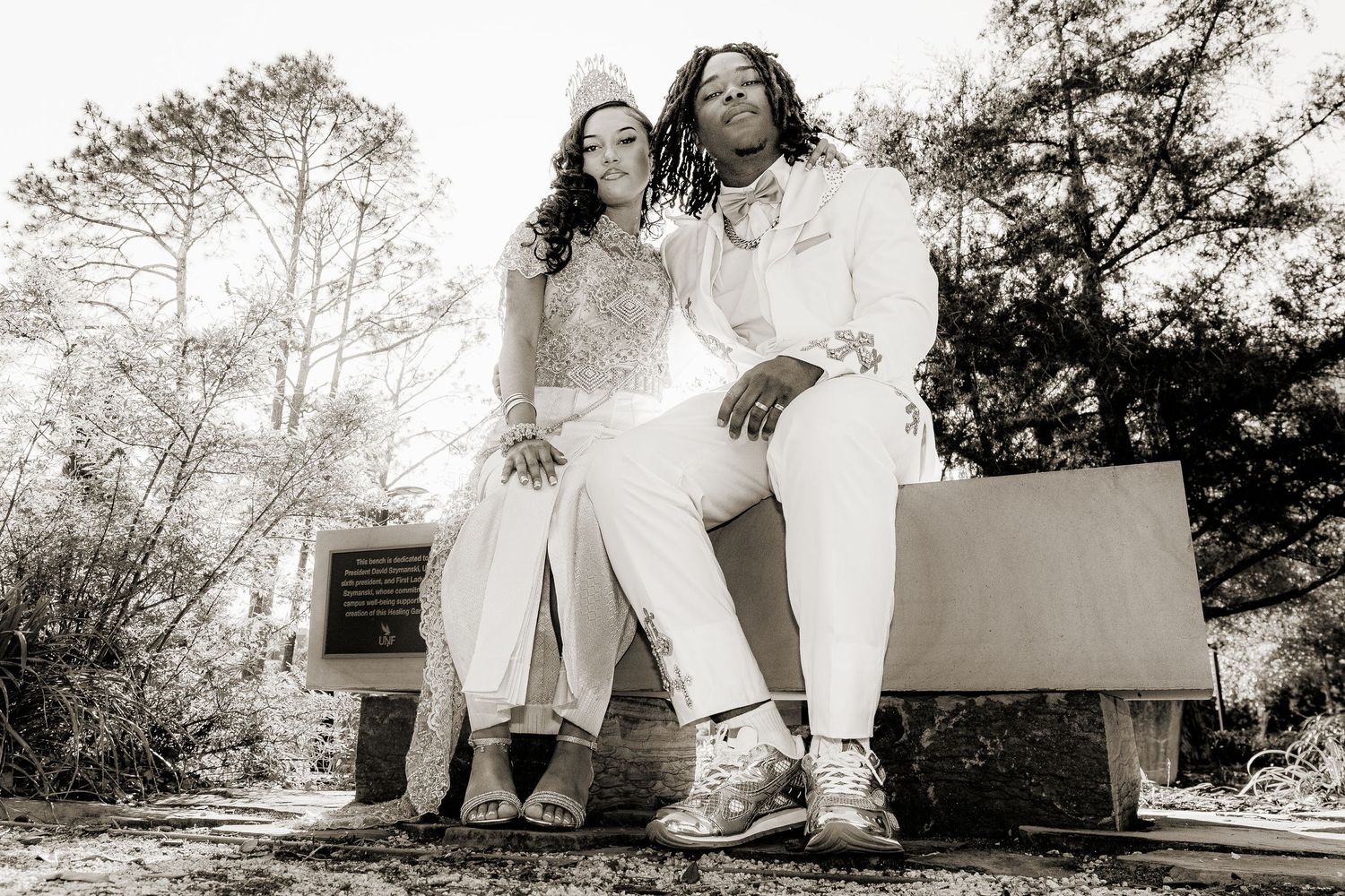 Couple in white outfits posing on a bench outdoors in a black and white wedding photo.
