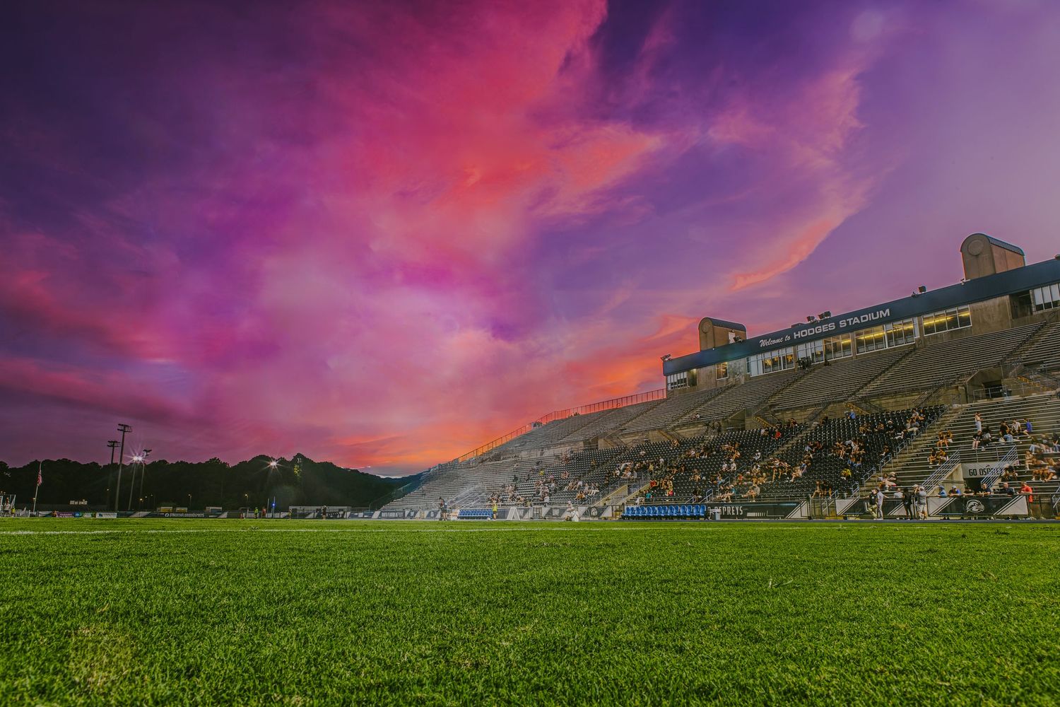 Dramatic purple and pink sunset sky over sports field with buildings in silhouette.