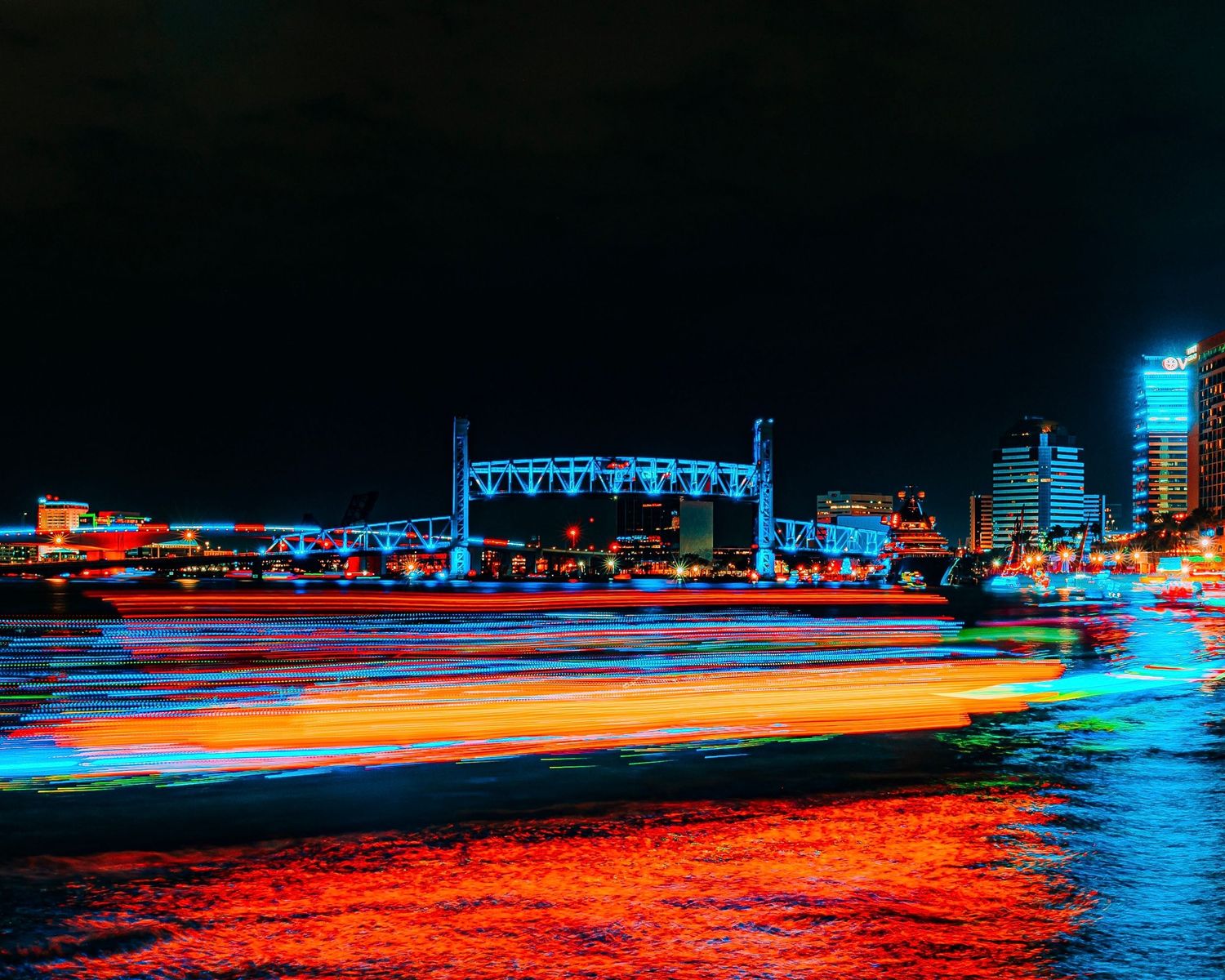 Long exposure cityscape with colorful light trails reflecting on water at night.