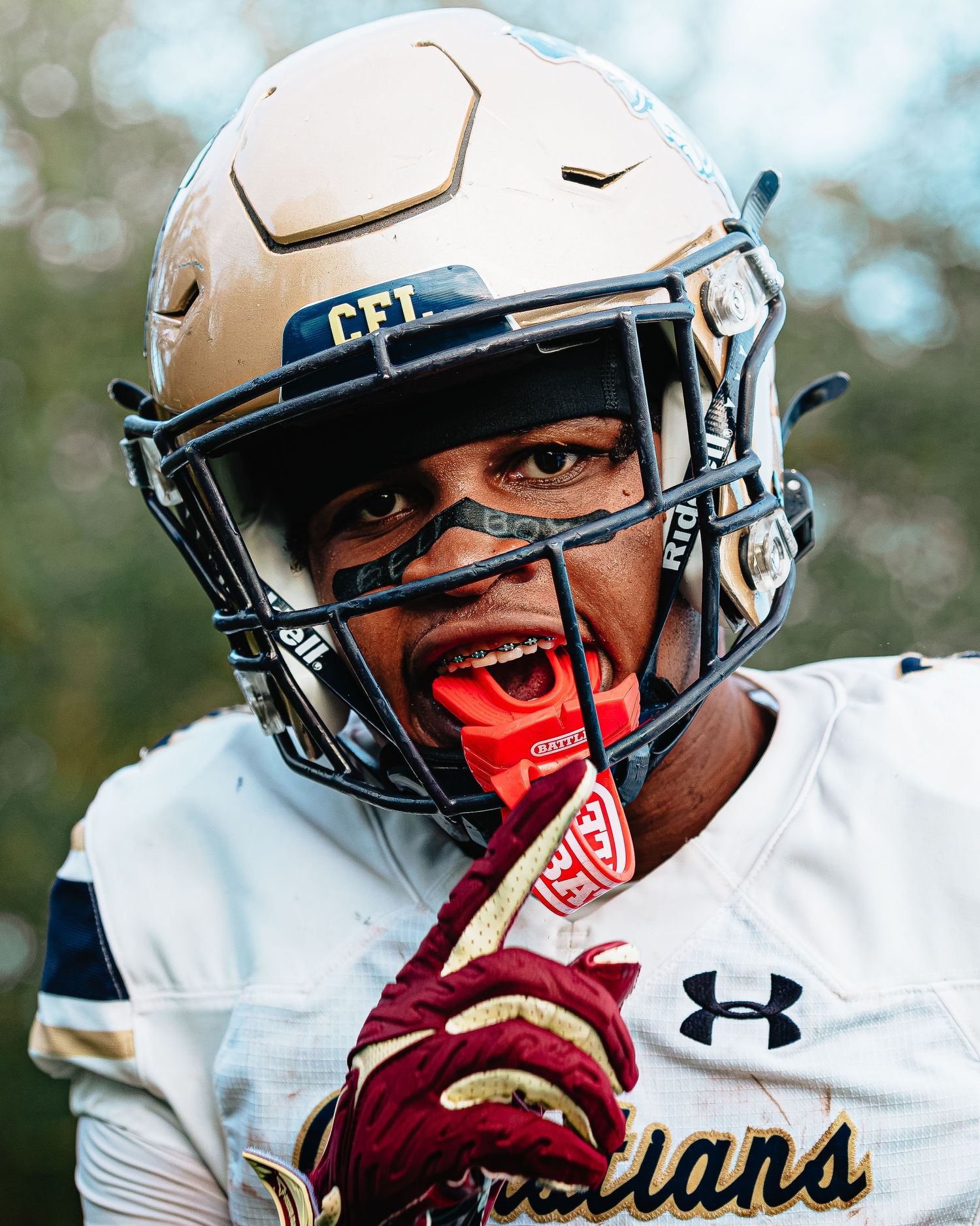 Football player in gold CFI helmet and Under Armour gear poses intensely on the field.