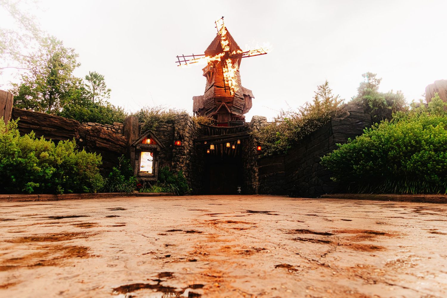 A whimsical windmill entrance with lanterns and lush greenery on a misty, rainy day at a fantasy-themed park.