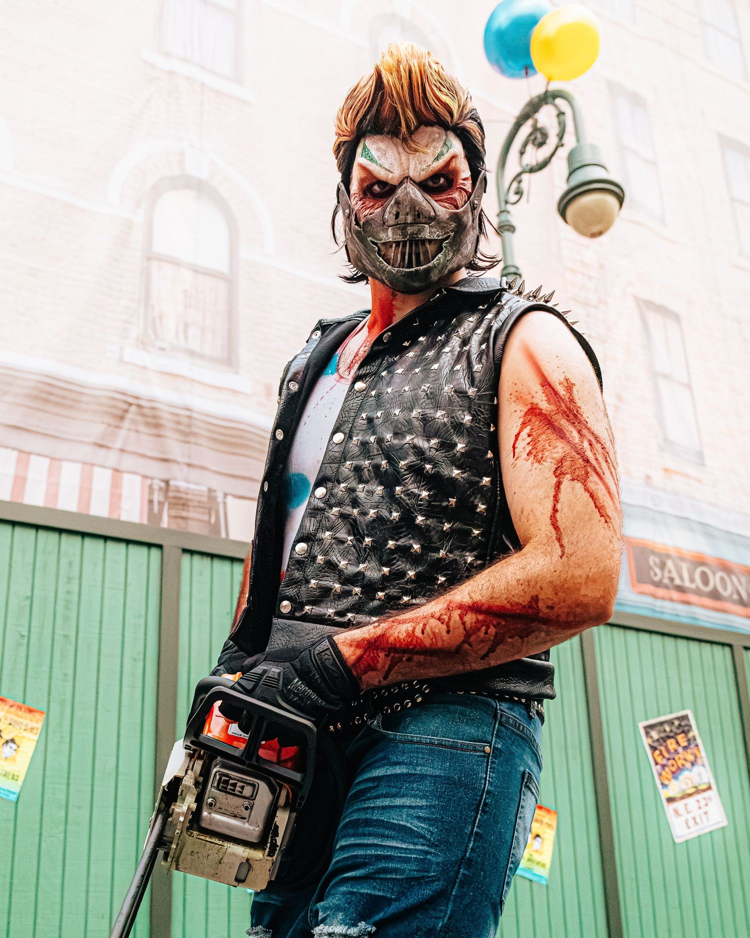 Person in horror costume with skull mask, bloody arm makeup, studded vest, and jeans posing on a street.