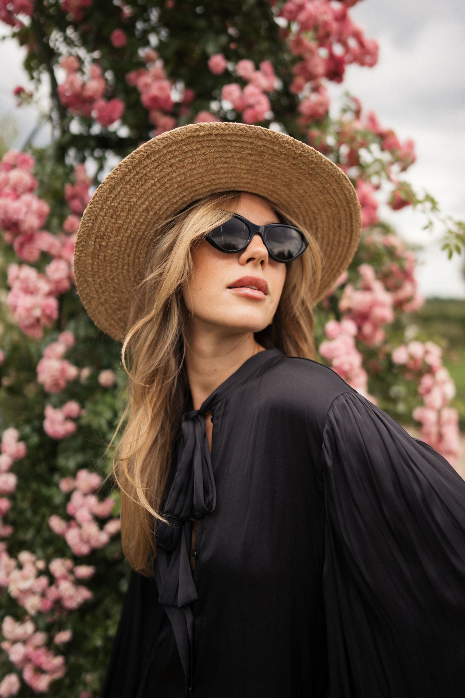 Woman wearing straw hat and sunglasses standing in front of flowering trees.
