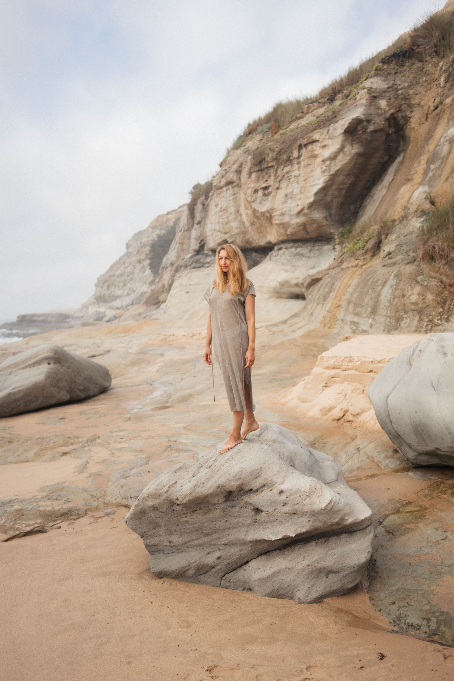 Woman standing on coastal rocks beneath sandstone cliff in soft natural light.